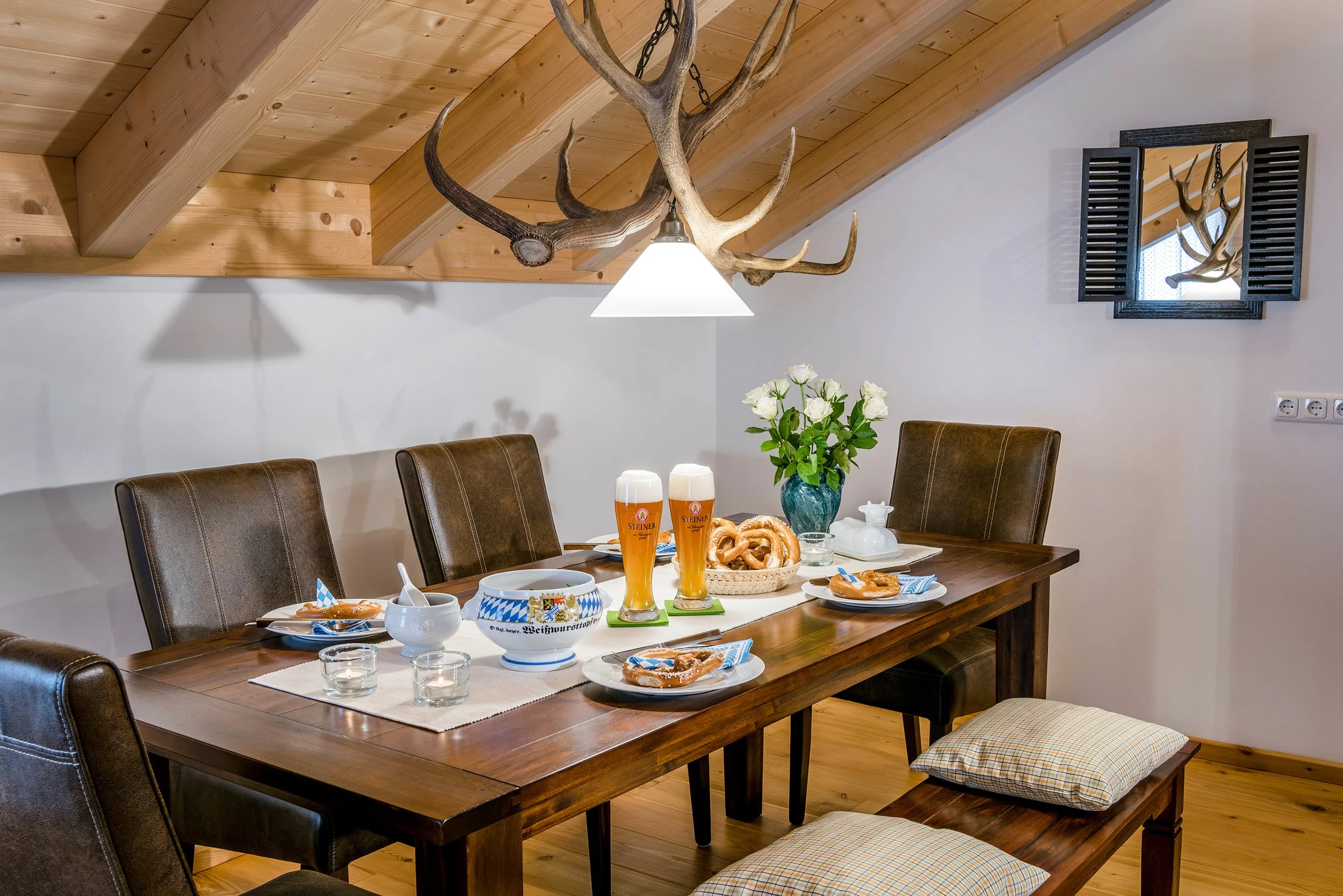 Ein festlich gedeckter Esstisch mit Bier, Brezeln, Töpfen, Blumen und Geschirr in einem Holzraum mit Wand- und Deckenbalken, einer Wandspiegel und einem Kronleuchter aus Ästen.