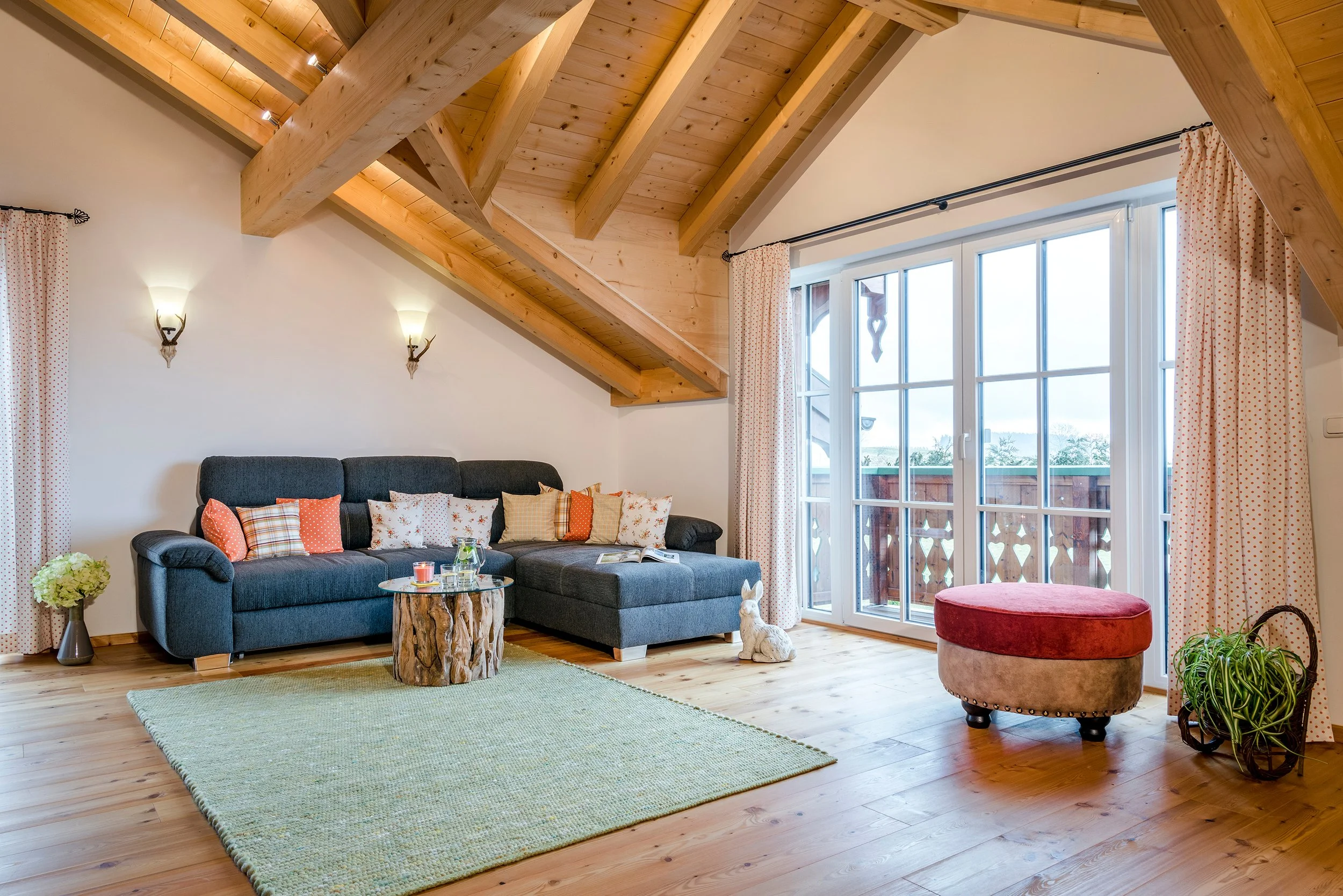 Ein Wohnzimmer mit einem grauen Sofa, einem kleinen Baumstümmertisch, einem grünen Teppich, einer roten Ottomane, Vorhängen mit roten Punkten, großen Fenstern und Holzbalken an der Decke.