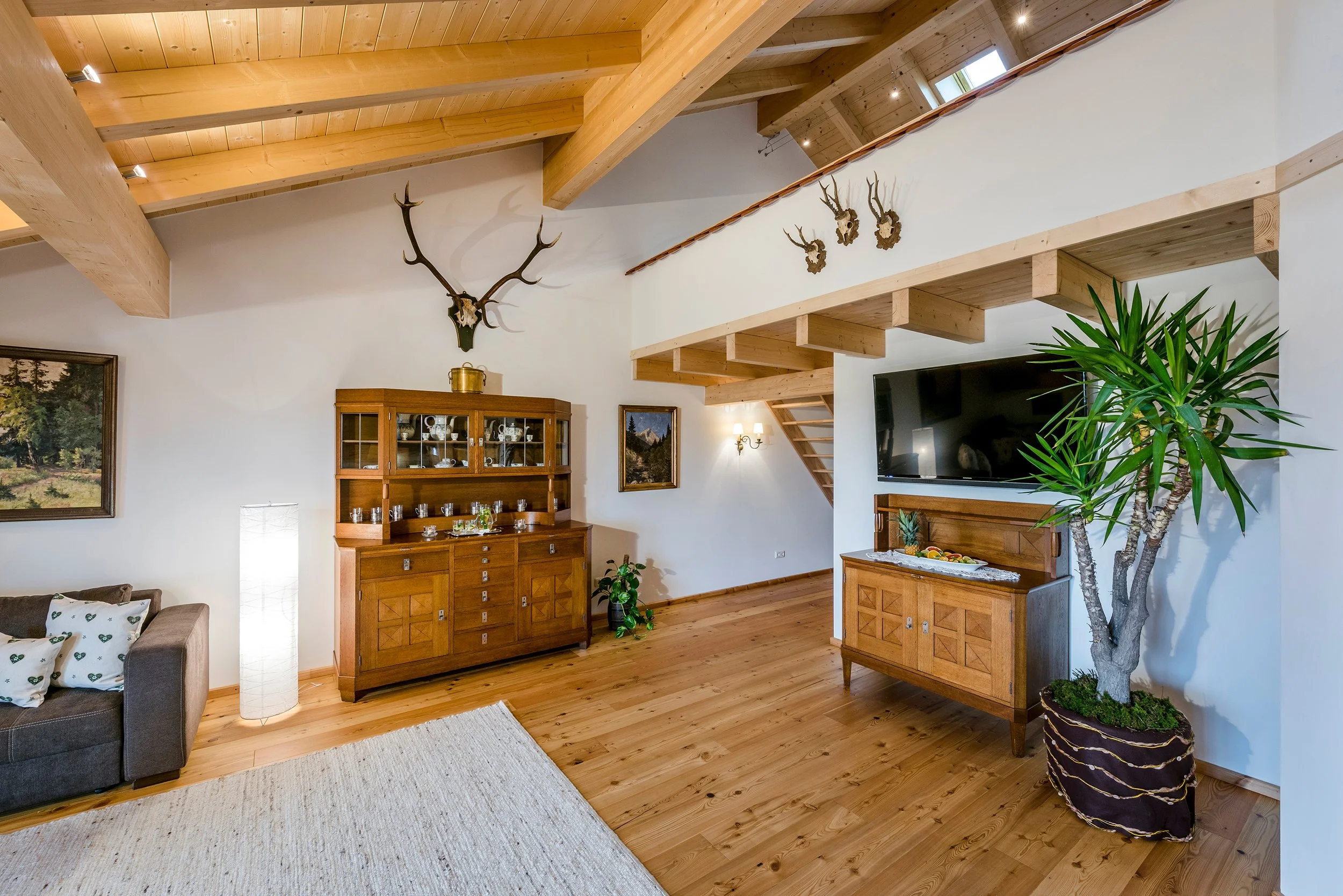 Rustikales Wohnzimmer mit Holzbalkendecke, Geweihen, Holzschrank, Fernseher, Treppe und großer Zimmerpflanze.