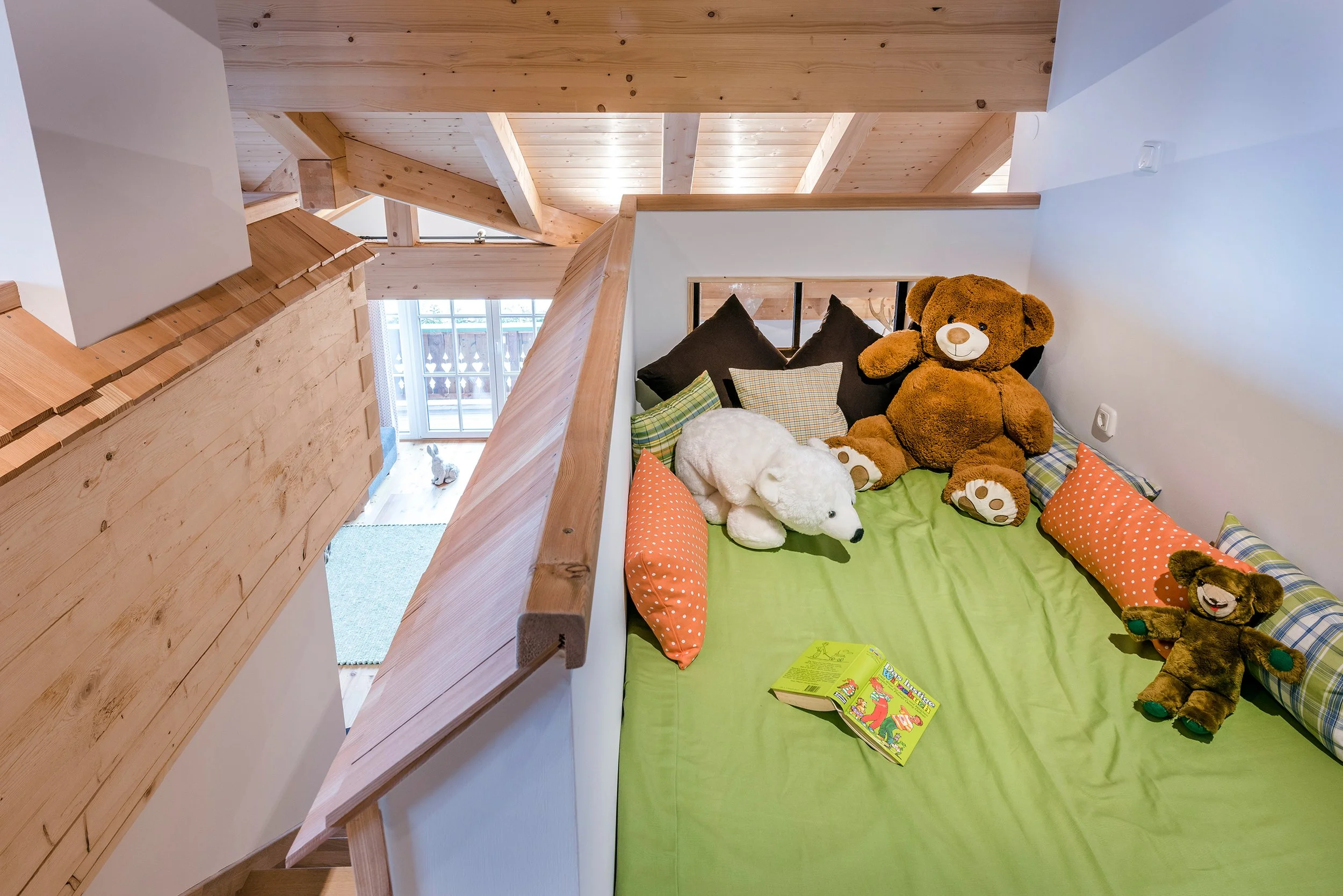 Gemütliches Spielecke auf einer Galerie mit grünem Bett, großem Teddybären, Stofftieren und Kinderbuch.