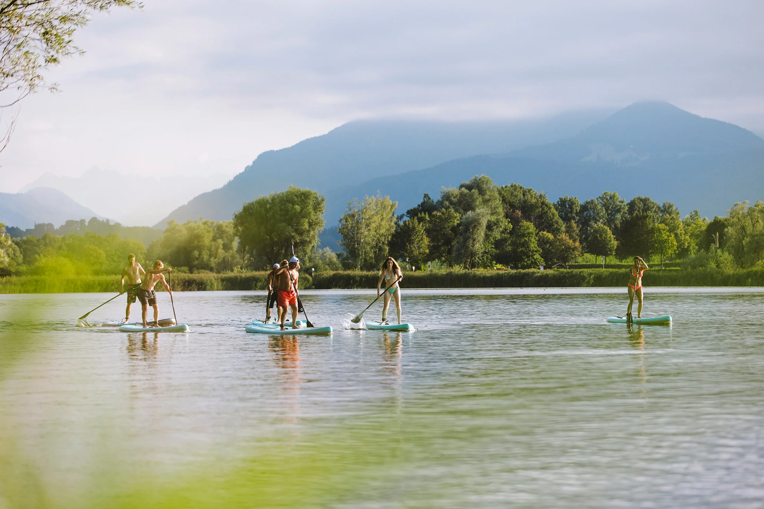 Menschen beim Stand-Up-Paddling auf einem See mit Bergen im Hintergrund, der See ist umgeben von grünen Bäumen.