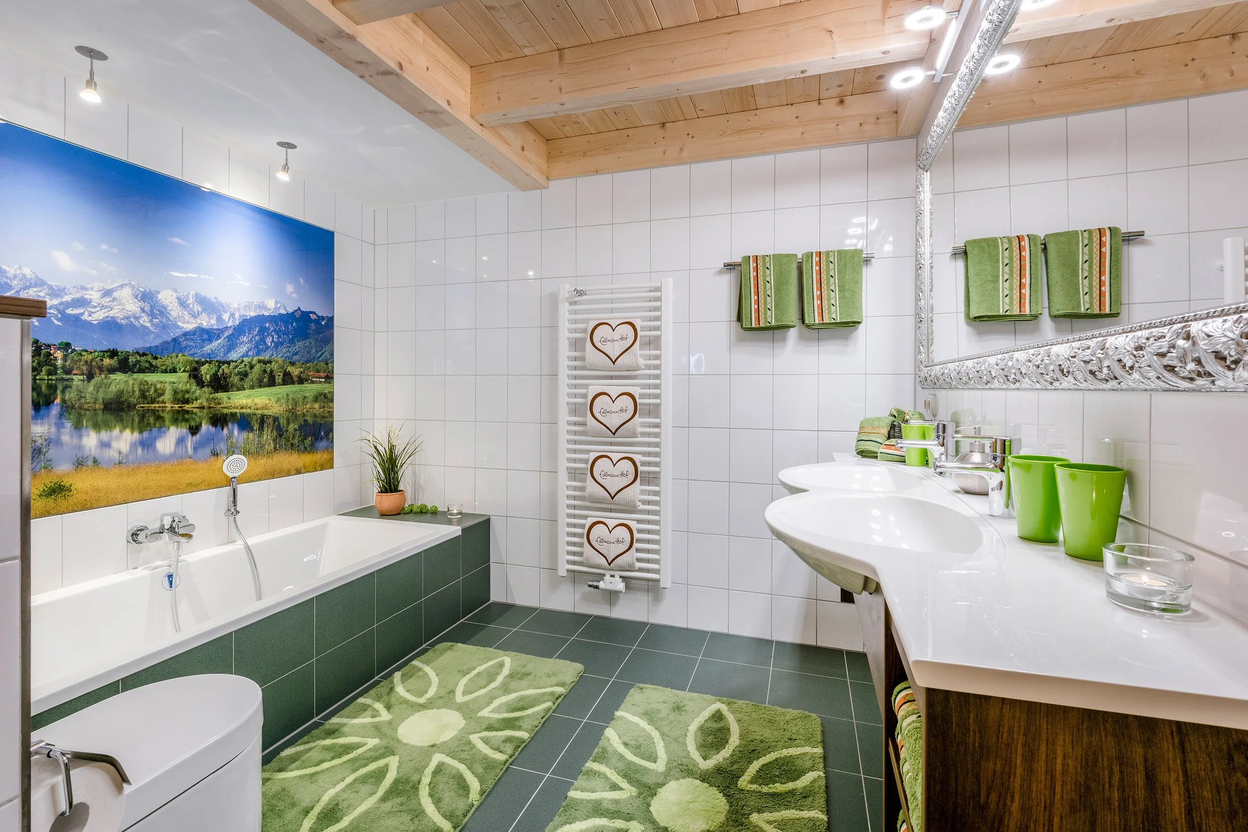 Badezimmer mit weißen Fliesen, grünen Handtüchern, großen Spiegel, Doppelwaschbecken, Badewanne mit Berglandschaft als Wandbild, grüne Badmatten, Pflanzen und grünen Geschirr.
