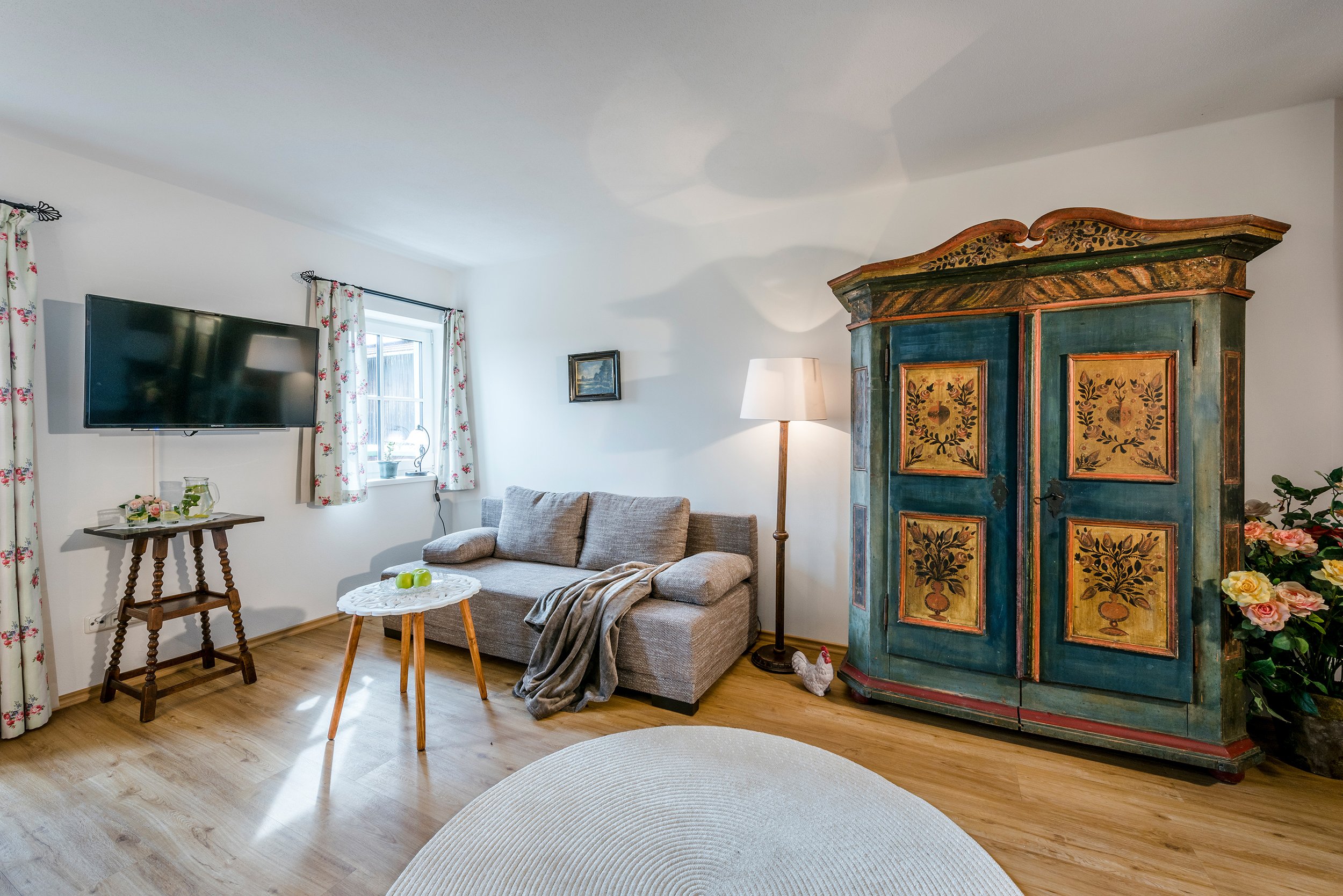 Ein gemütliches Wohnzimmer mit einem grauen Sofa, einem kleinen runden Couchtisch mit Äpfeln, einem alten verkleideten Schrank, einem Tisch mit Blumen, einem Fernseher, einem Wandbild, einer Stehlampe, und hohen Fenstern mit weißen Vorhängen mit roten Blumenmuster.