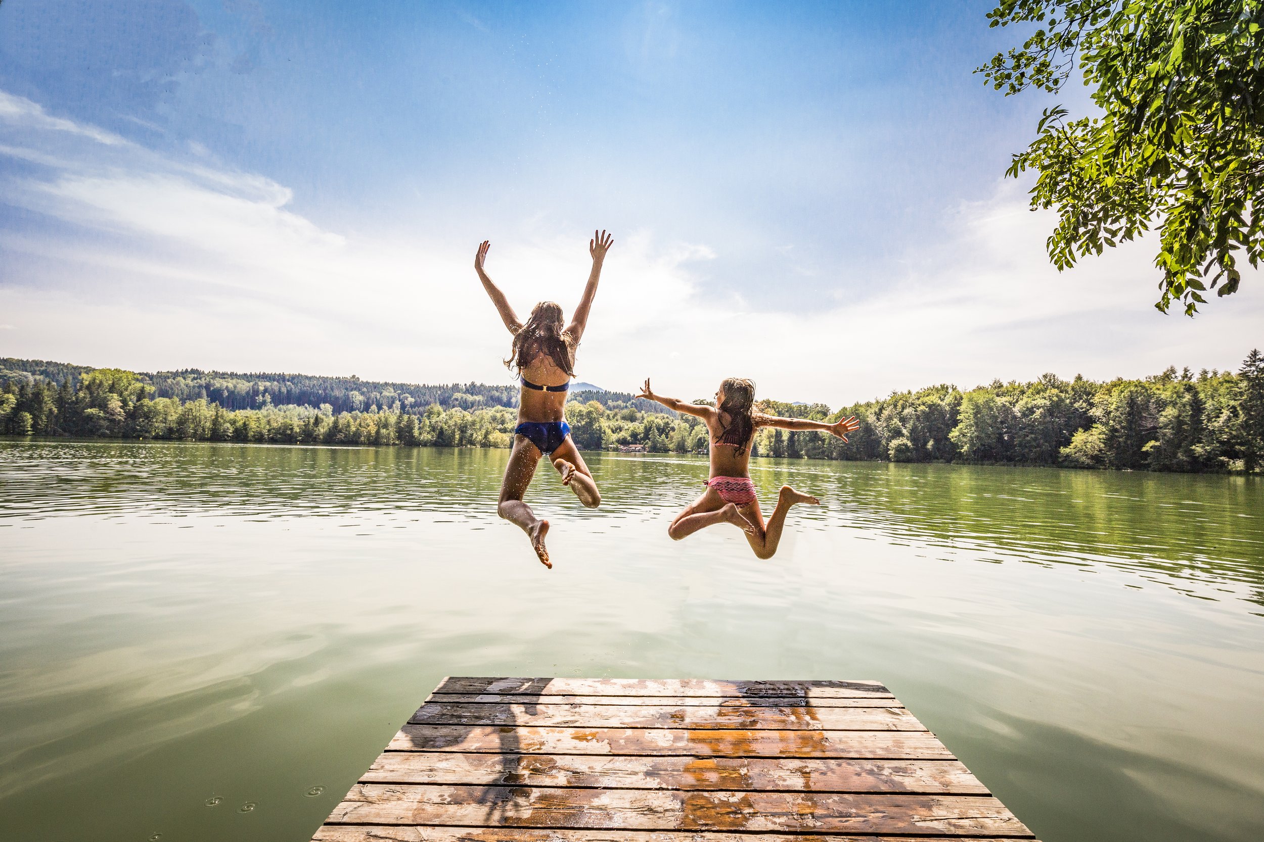 Zwei Kinder springen von einem Steg in einen See bei Sonnenschein, umgeben von grünen Bäumen.
