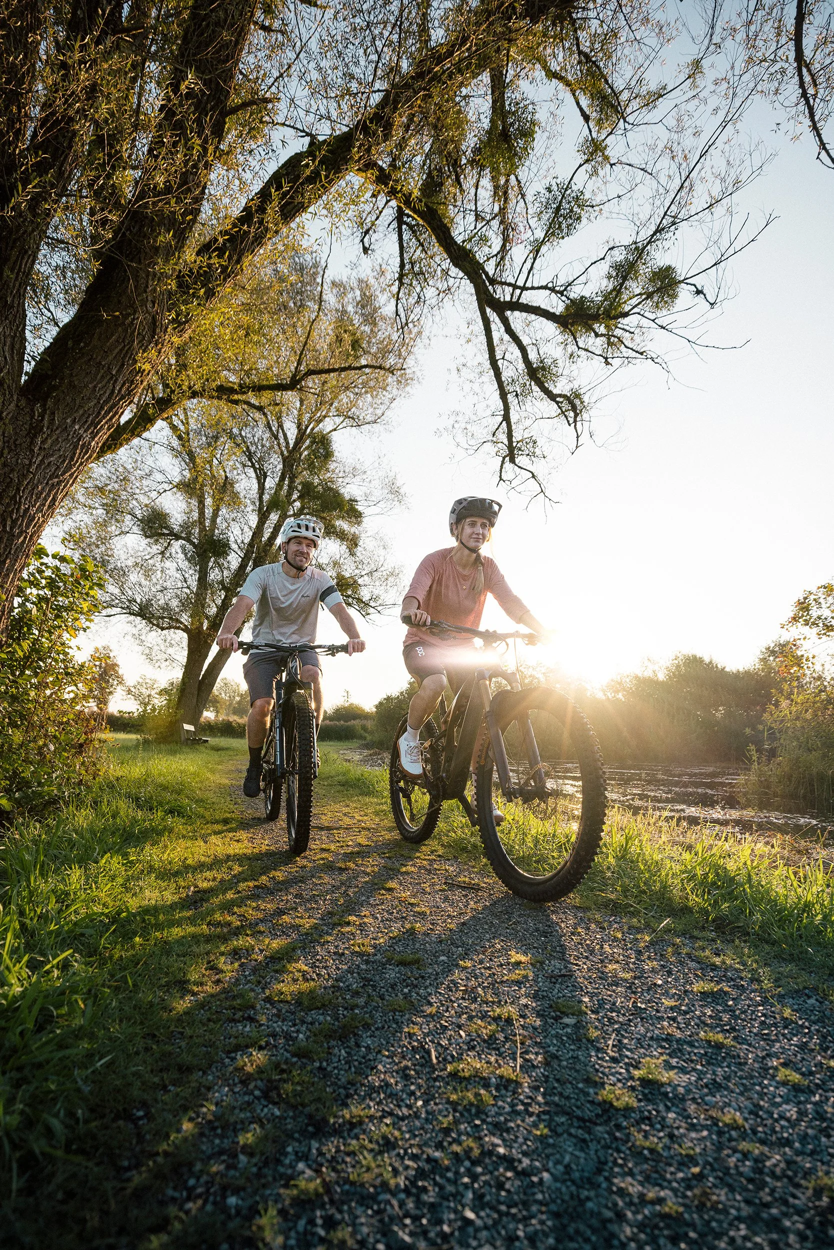 Zwei Menschen fahren mit E-Bikes auf einem Pfad entlang eines Flusses bei Sonnenuntergang. Es sind Bäume im Hintergrund und das Licht ist warm.