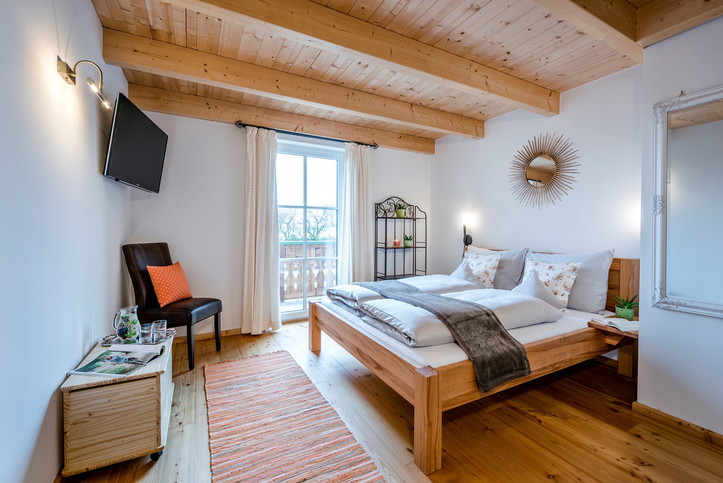 Schlafzimmer mit Holzbalkendecke, weißer Wand, Bett mit weißer Bettwäsche und grauem Überwurf, Spiegel mit Sonnenstrahlenrahmen, kleine Regal mit Pflanzen, Fenster mit weißen Vorhängen, Sitzbank mit Zeitschriften, Teppich auf Holzboden, Flachbildfernseher