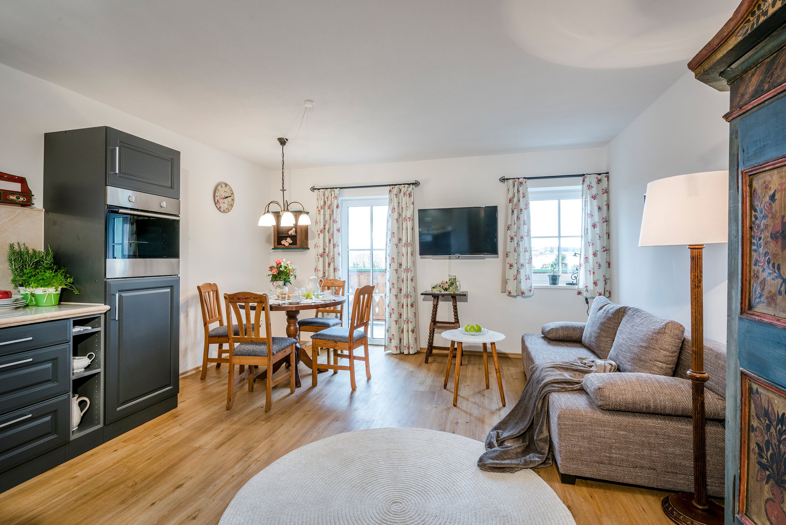 Helles Wohn- und Esszimmer der Ferienwohnung Rosengarten, einem großen Fernseher, einem grauen Sofa, einem runden Teppich, einem Couchtisch, einem Essbereich mit Holzstühlen, bunten Vorhängen, einem Schrank und einer Wanduhr.
