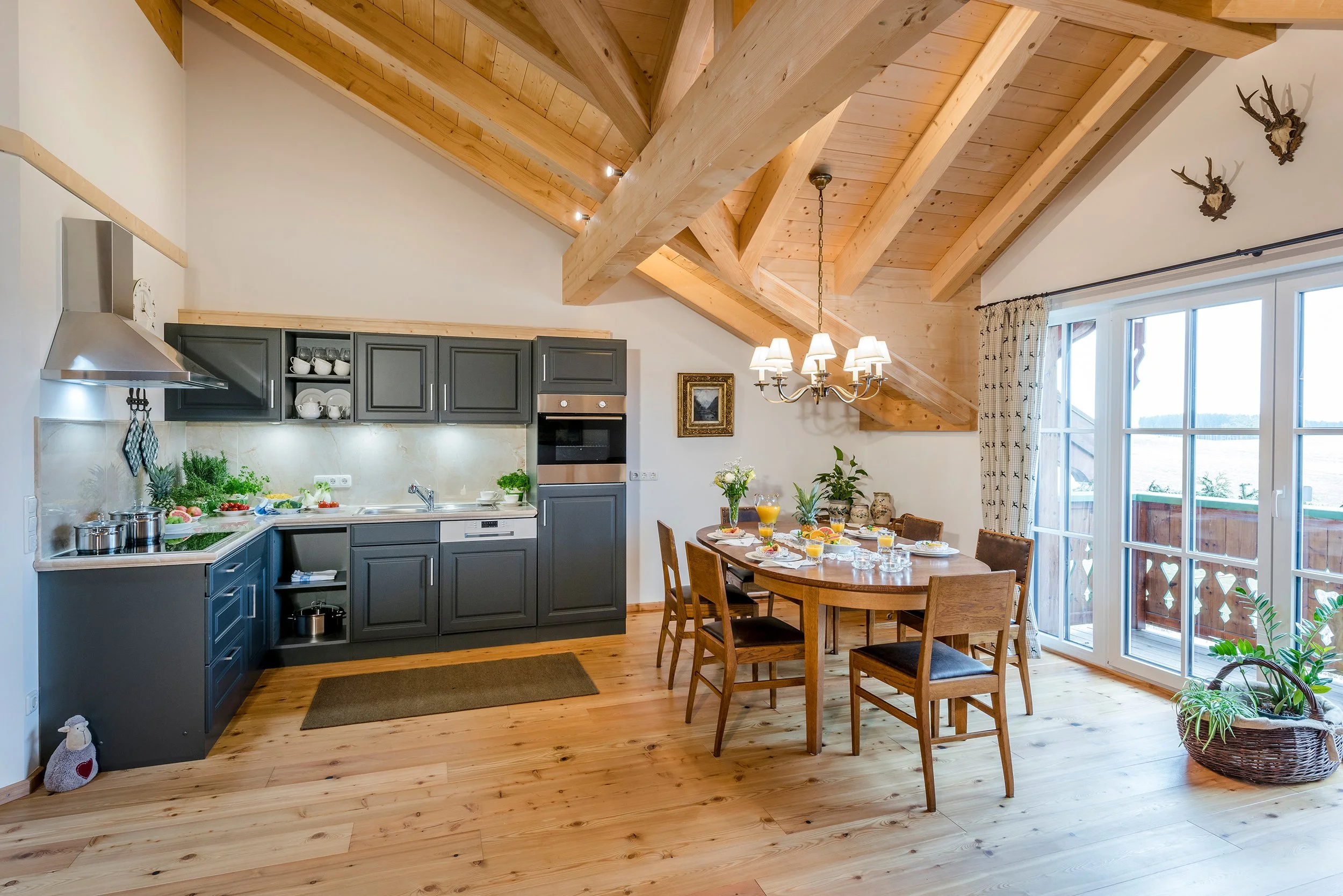 Heller Wohn- und Essbereich mit Holzbalken, Esstisch mit Frühstück, große Fenster mit Gardinen, Küchenbereich mit dunklen Schränken