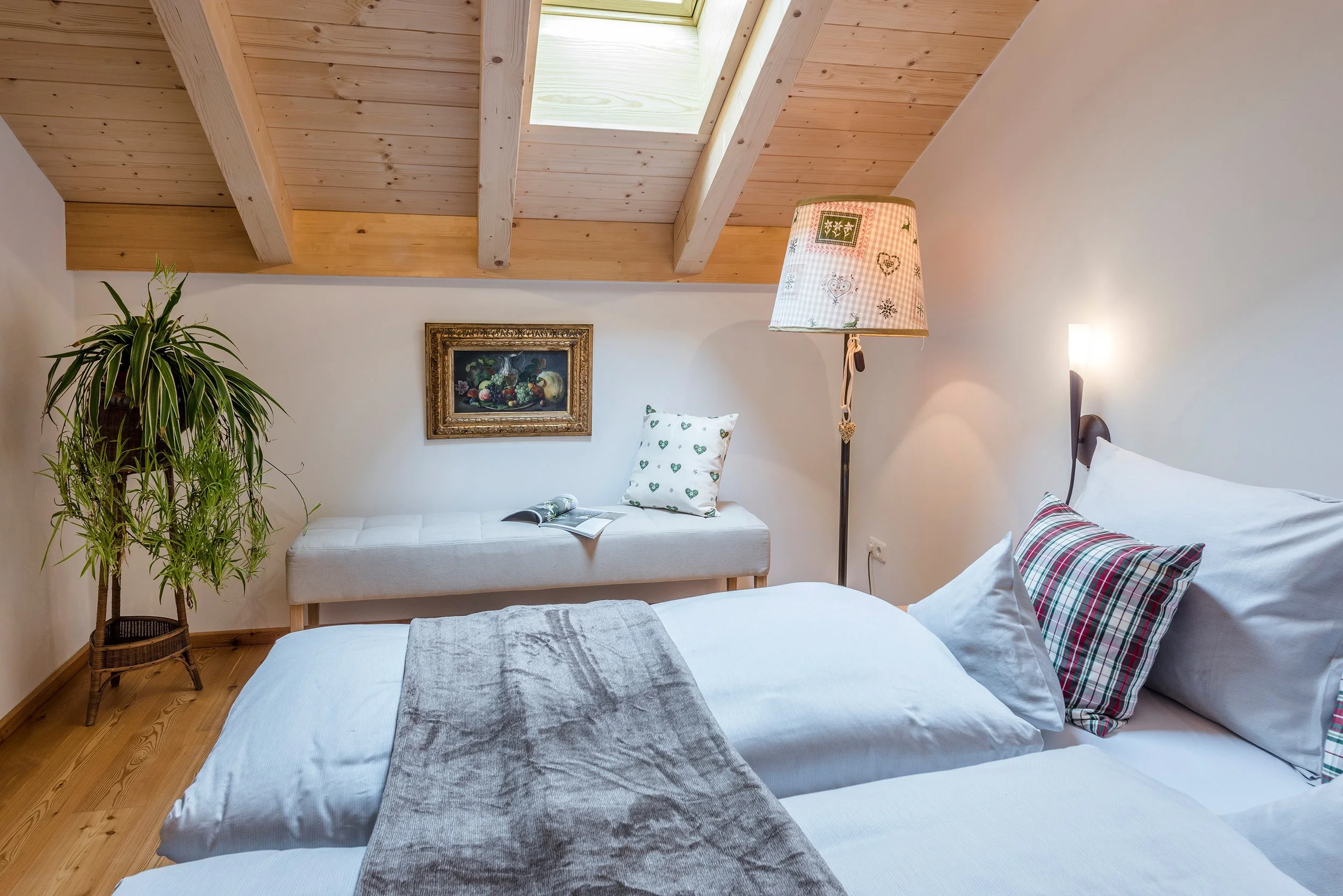 Ein Schlafzimmer mit weißer Bettwäsche, Kissen mit grünen Herzmustern, einer Leselampe im Landhausstil, einer Pflanze, einem gerahmten Stillleben-Bild und einem Dachfenster in einer Holzdecke.
