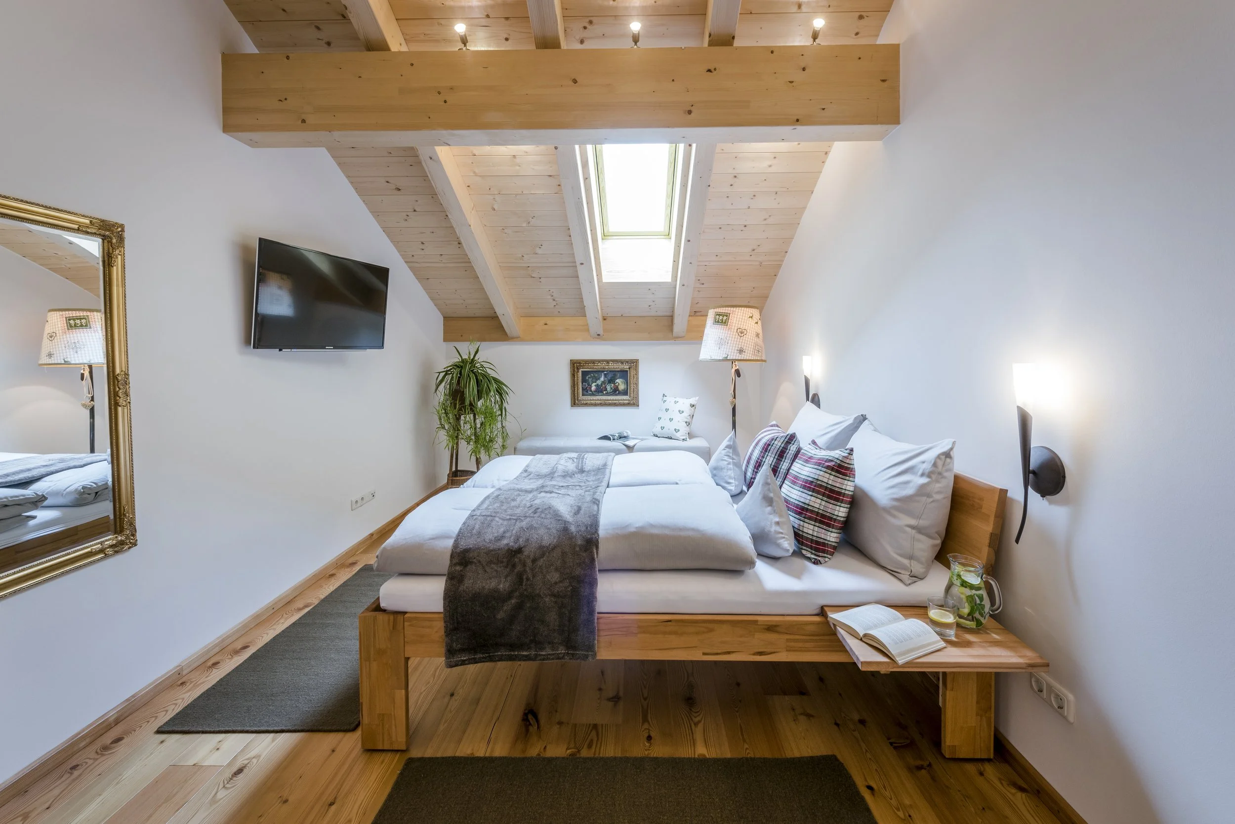Ein Schlafzimmer mit Holzdecke, einem Bett mit weißer Bettwäsche und mehreren Kissen, einem kleinen Tisch mit einem offenen Buch und Getränken, einer Stehlampe, einem Fernseher an der Wand, einem Spiegel mit goldenem Rahmen, Pflanzen und Skulpturen im Hintergrund.