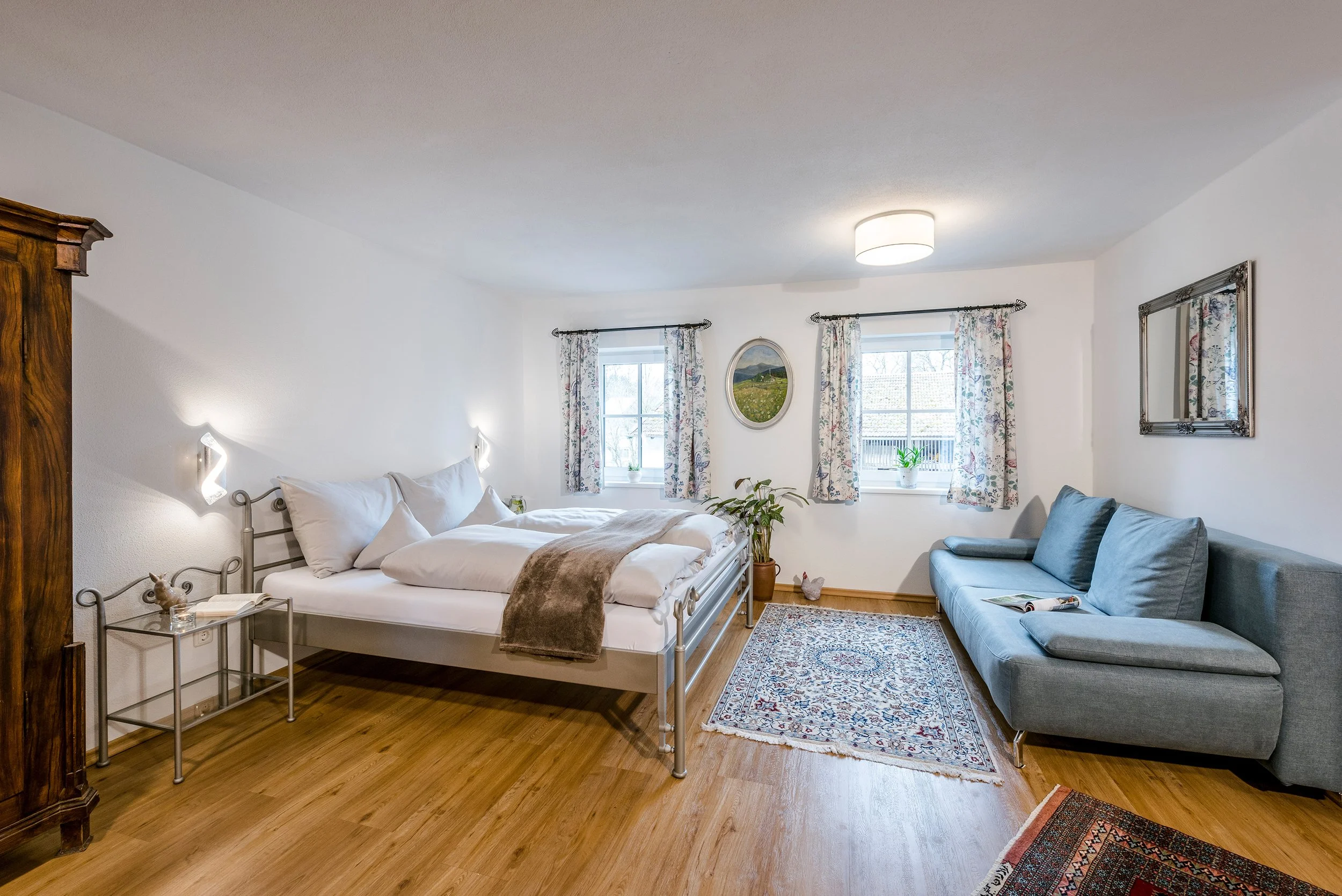 Gemütliches Wohn- und Esszimmer der Ferienwohnung Lindenblick mit einem Bett, Bettwäsche und Kissen, Holzboden, weißen Wänden, einem Teppich, einem grauen Sofa, zwei Fenstern mit bunten Gardinen, einem Spiegel, Pflanzen und dekorativem Wandbild.