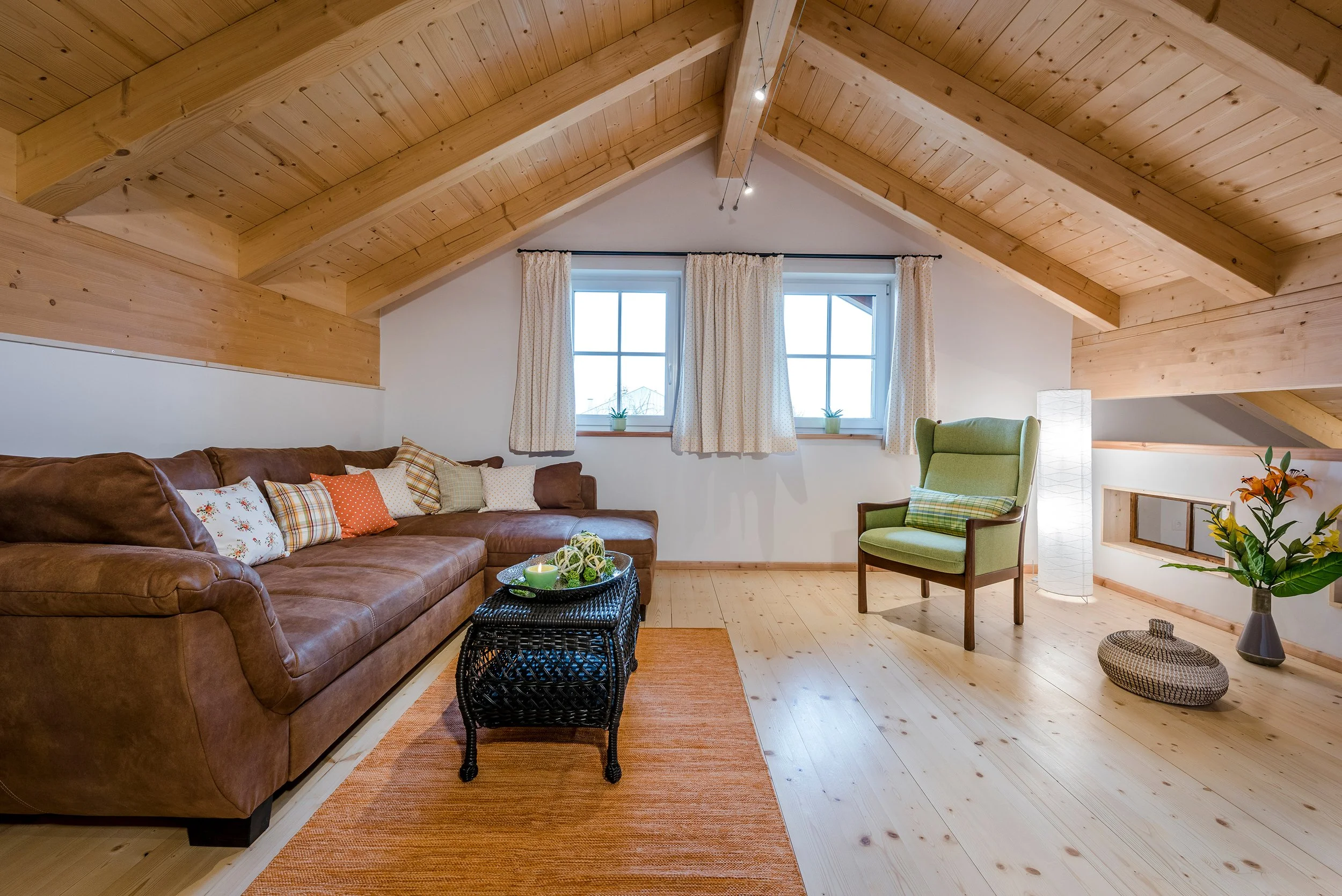 Galerie der Ferienwohnung Allpenglück mit Holzbalkendecke, zwei Fenster mit weißen Vorhängen, braunes Sofa mit bunten Kissen, grüner Sessel, Tisch mit Dekoration, Laminatboden und Stehleuchte.