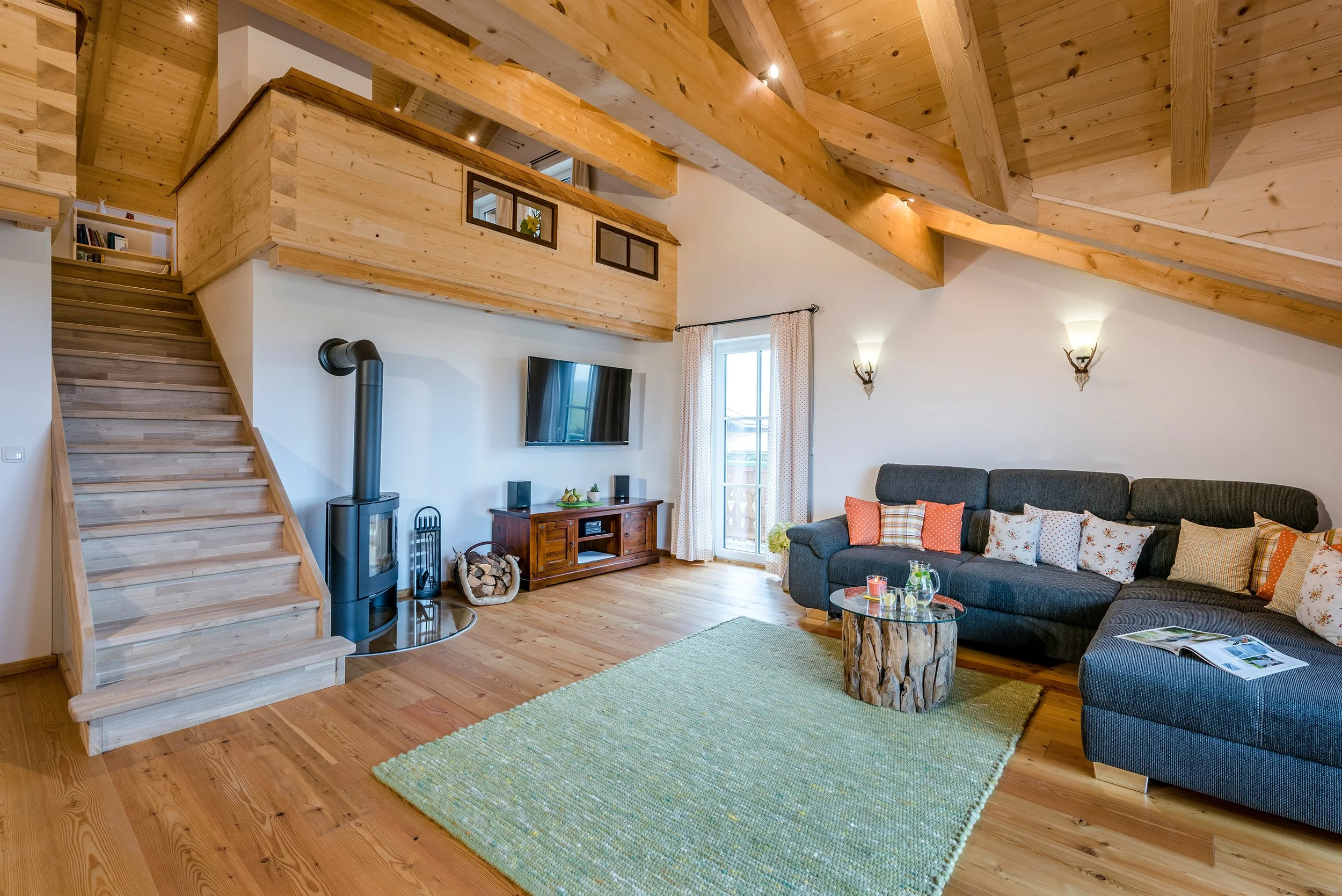 Innenansicht eines gemütlichen Wohnzimmers mit Holzbalken an der Decke, einem grauen Sofa mit verschiedenen Kissen, einem runden Couchtisch aus Holz, einem Kamin, einem Fernseher und einer gläsernen Tür, die auf einen Balkon führt.