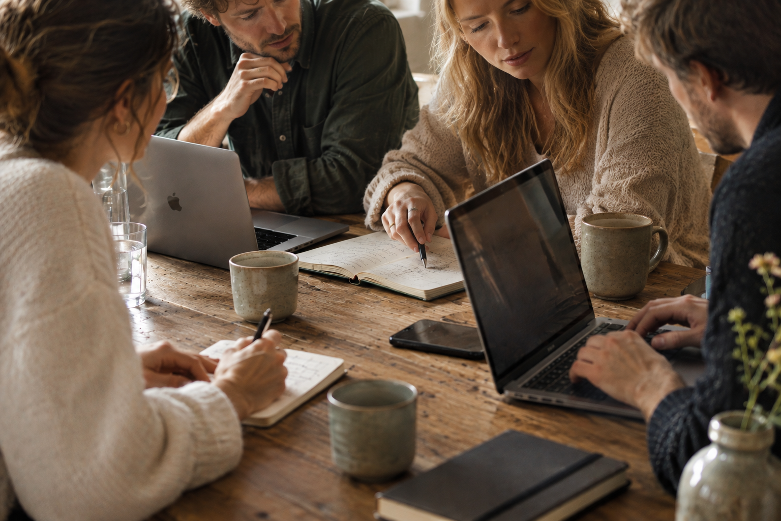 Fünf Menschen sitzen um einen Holztisch, diskutieren, schreiben in Notizbücher, arbeiten an Laptops, haben Kaffeetassen vor sich und schauen auf ein Notizbuch.
