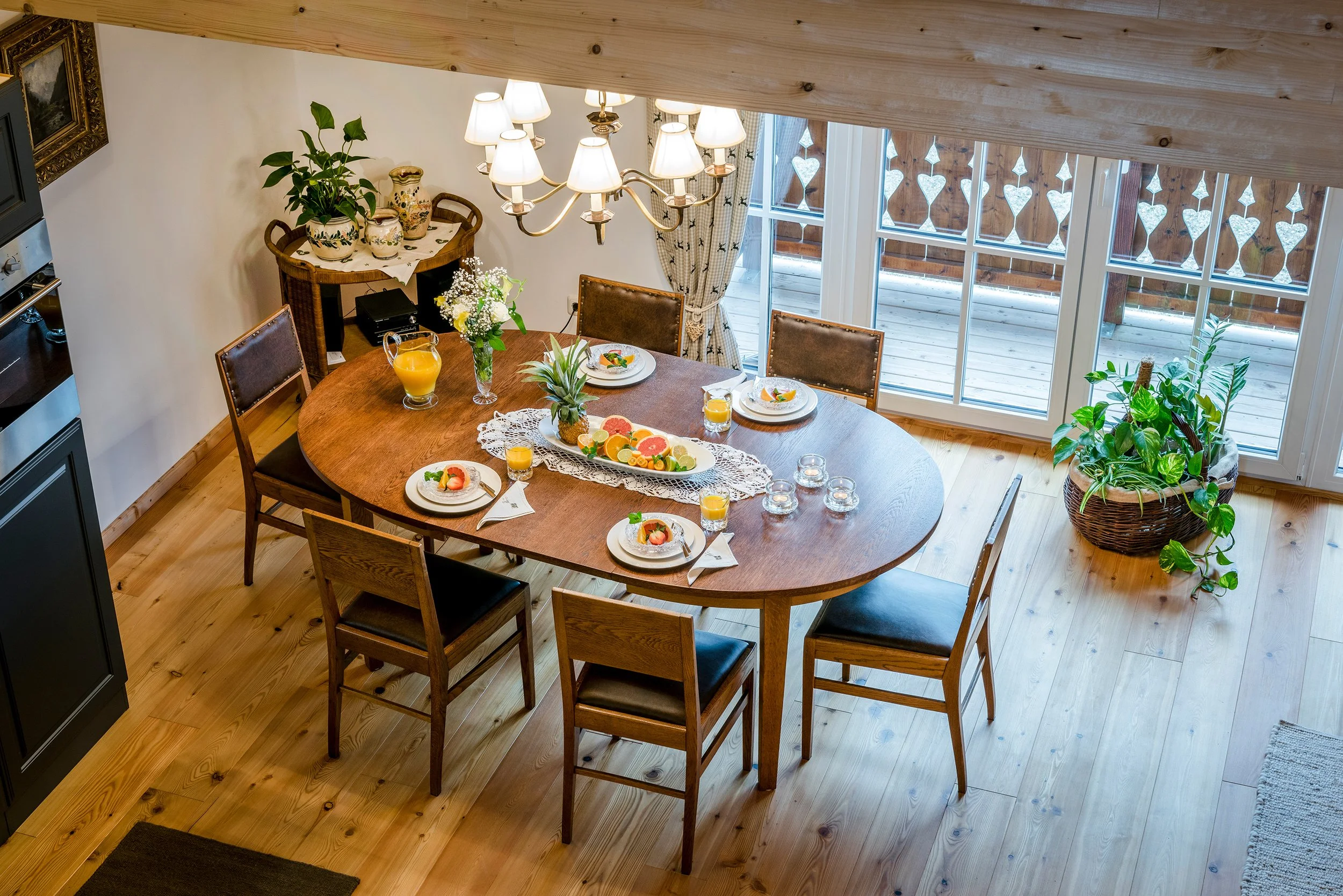 Ein Holztisch mit Frühstück, darunter ein Teppich. Der Tisch ist mit Orangensaft, Früchten und Tellern mit Gekochtem gedeckt. Es gibt mehrere Stühle, eine große Fensterfront mit Herz- und Diamantförmchen dekoriert, Stehlampe und Pflanzen im Raum.