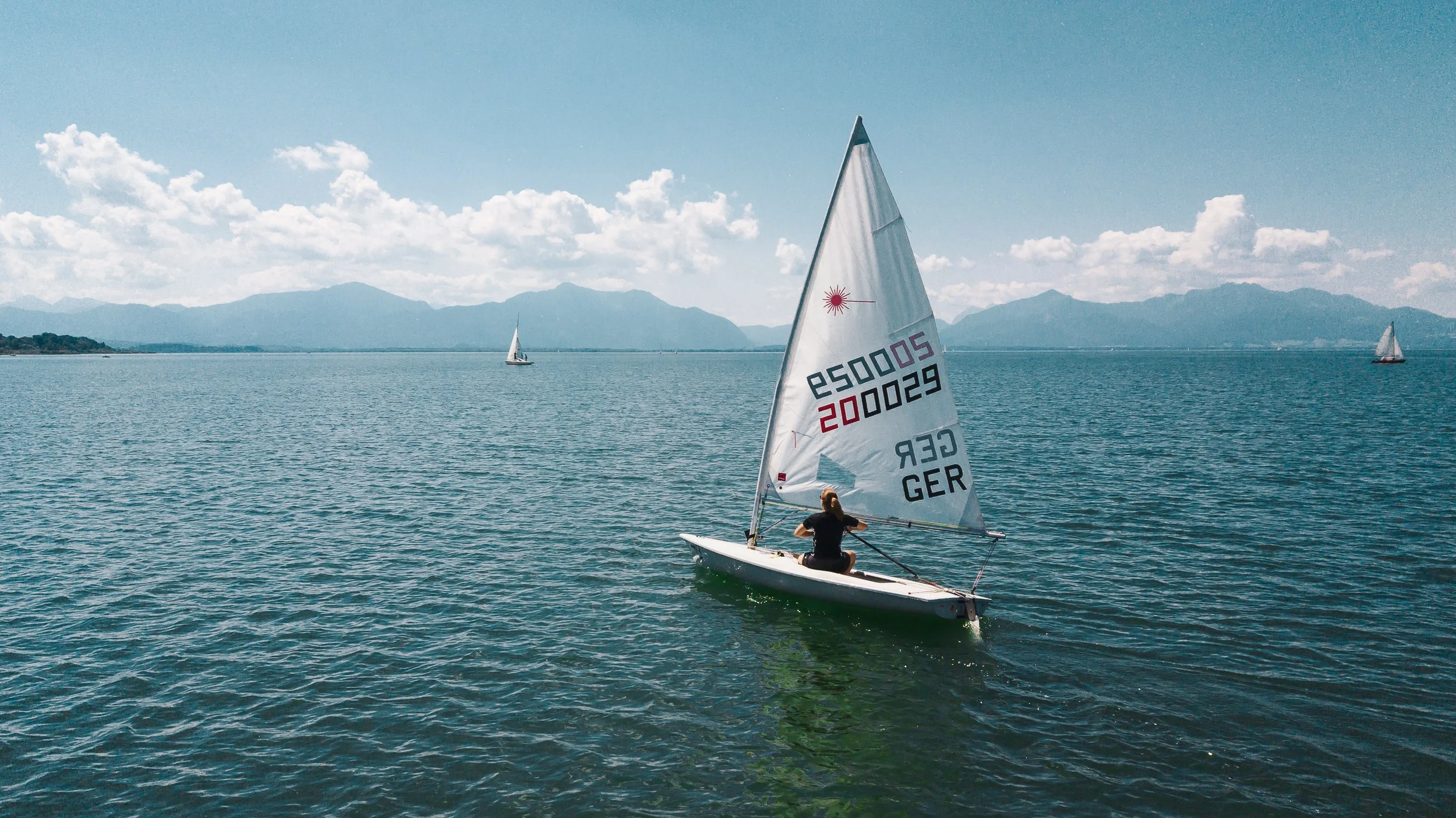 Eine Person segelt auf einem See mit mehreren Segelbooten im Hintergrund. Es ist ein klarer Tag mit bewölktem Himmel und Bergen im Hintergrund.