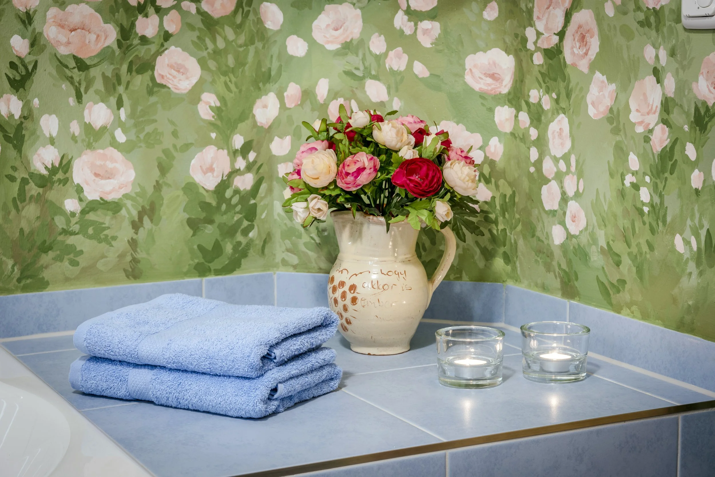 Blumenstrauß mit rosa, weißen und roten Blumen in einer Keramikvase auf einem Badezimmerwaschbecken, neben einem blauen Handtuch und drei Glas-Kerzenhaltern mit Kerzen, vor einer grünen Wand mit rosa Blumenmuster.