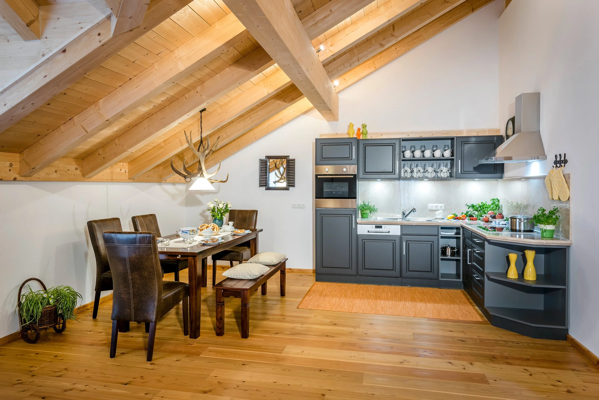Offene Küche im rustikalen Stil mit Schränken, Holzdecke mit sichtbaren Balken, Esstisch mit Stühlen, Pflanzen und Dekorationen auf der Arbeitsplatte.