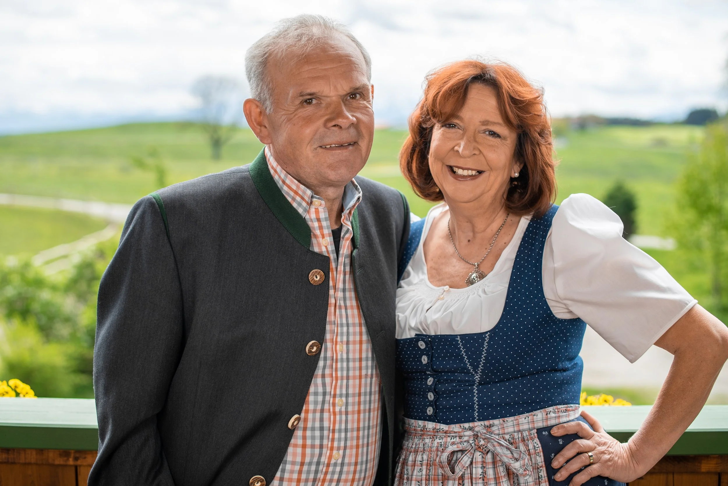 Ein älteres Paar in traditioneller deutscher Kleidung, lächelnd mit einer grünen Landschaft im Hintergrund.