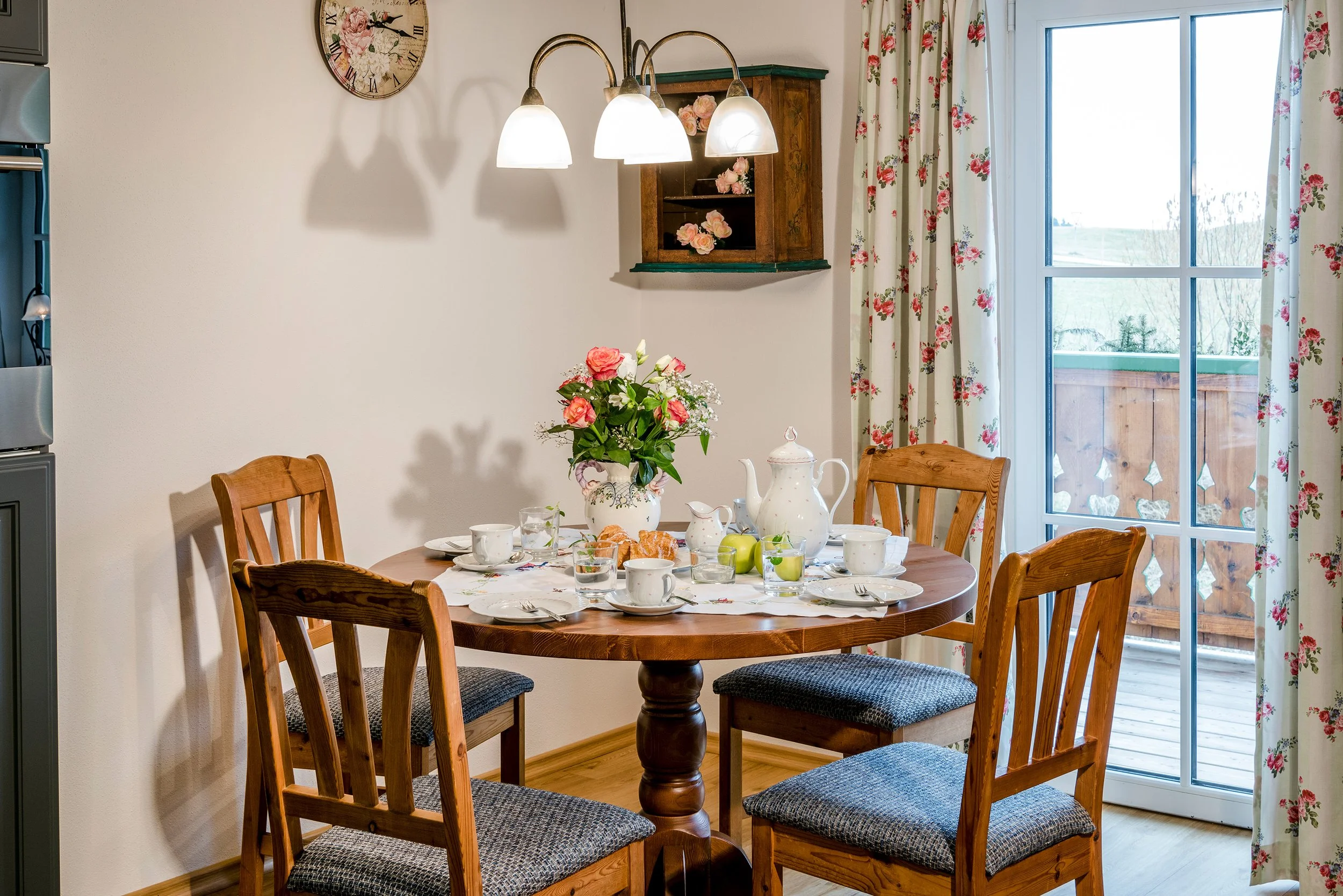 Gedeckter Esstisch der Ferienwohnung Rosengarten mit Blumen, Geschirr und Besteck, im Hintergrund eine Fensterfront mit blumengeschmückten Vorhängen und ein Holzschrank, an der Wand eine Uhr und eine Lampe.