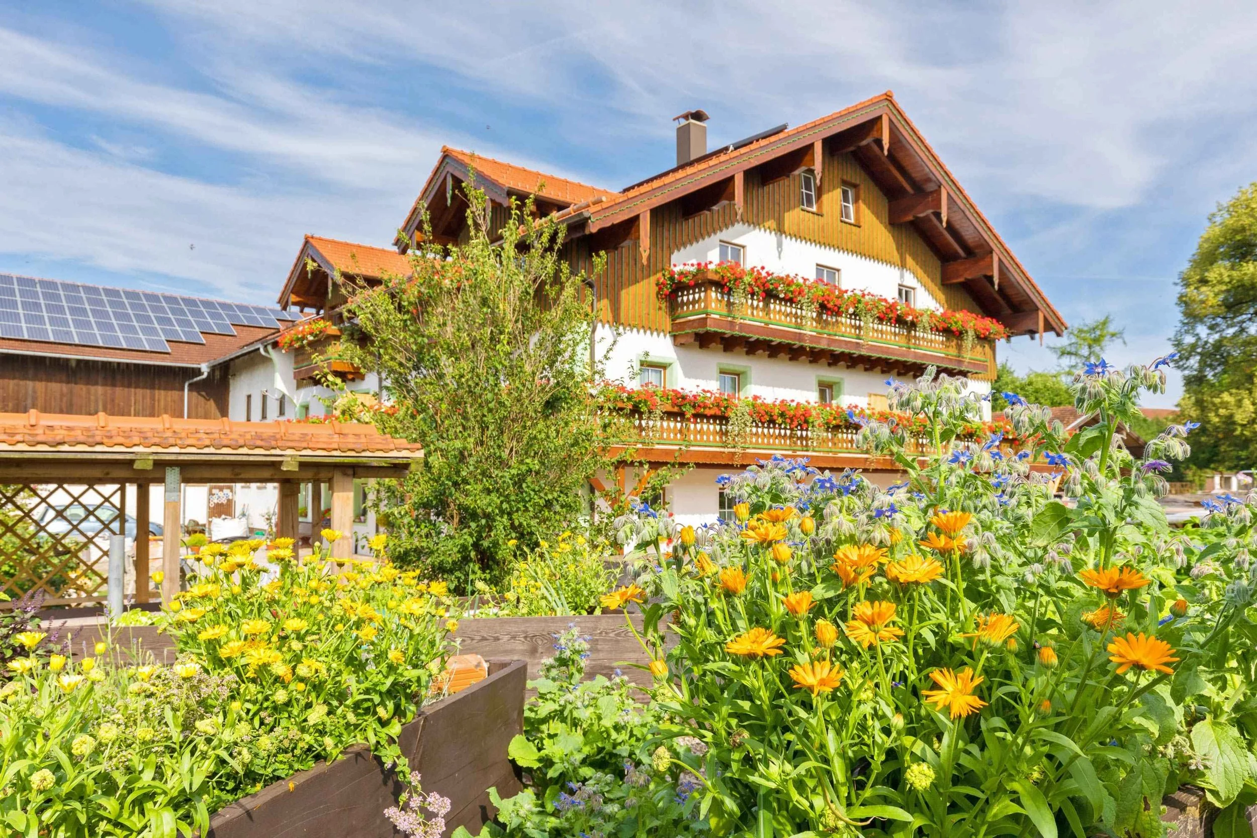 Ein mehrstöckiges Haus mit Holzwänden und roten Dachziegeln, umgeben von bunten Blumen und Bäumen im Garten, unter blauem Himmel.