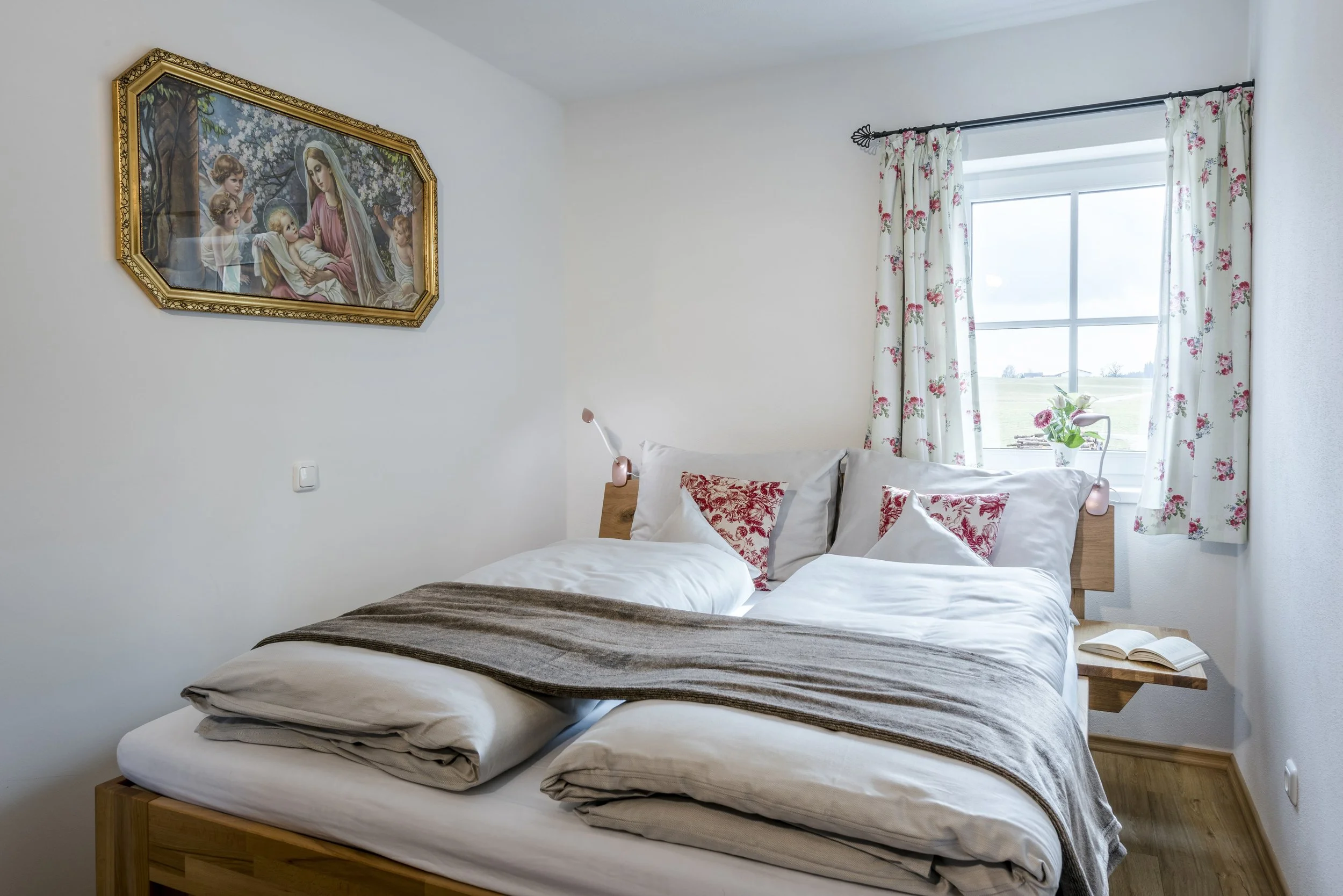 Schlafzimmer der Ferienwohnung Rosengarten mit weißen Wänden, einem Holzbett, weißen Bettwäsche, roten Kissen, einem Fenster mit Vorhängen und einer Blume auf der Fensterbank. An der Wand hängt ein Bild.