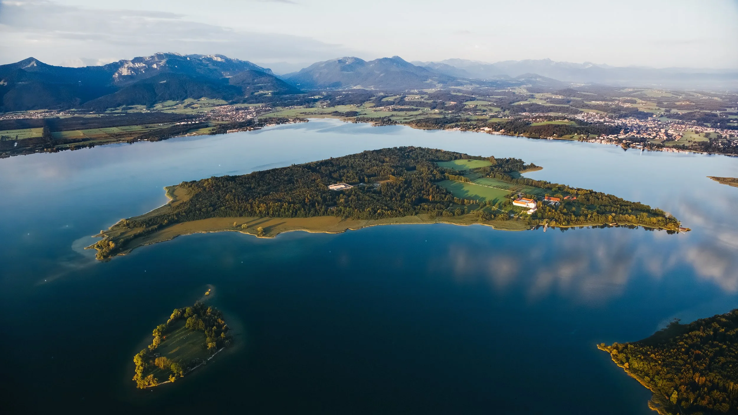 Luftaufnahme eines Sees mit einer kleinen, bewaldeten Insel in der Mitte, umgeben von flacher Landschaft und Bergen im Hintergrund.