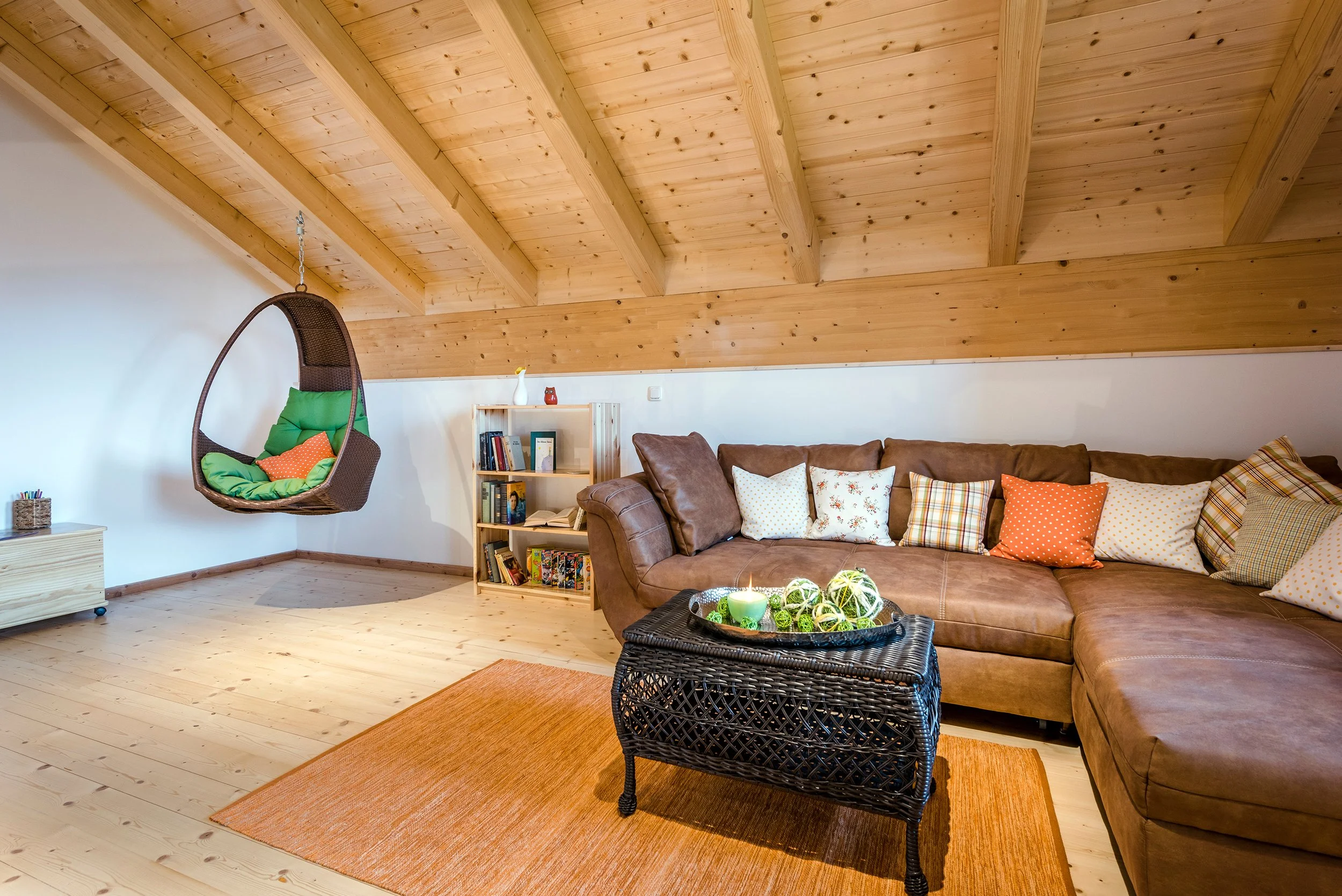 Galerie mit Holzbalkendecke, einem braunen Ledersofa, bunten Kissen, einem schwarzen Couchtisch mit dekorativen Kugeln, einem Bücherregal, einer Hängeschaukel mit Kissen und einem weißen Wandhaken.