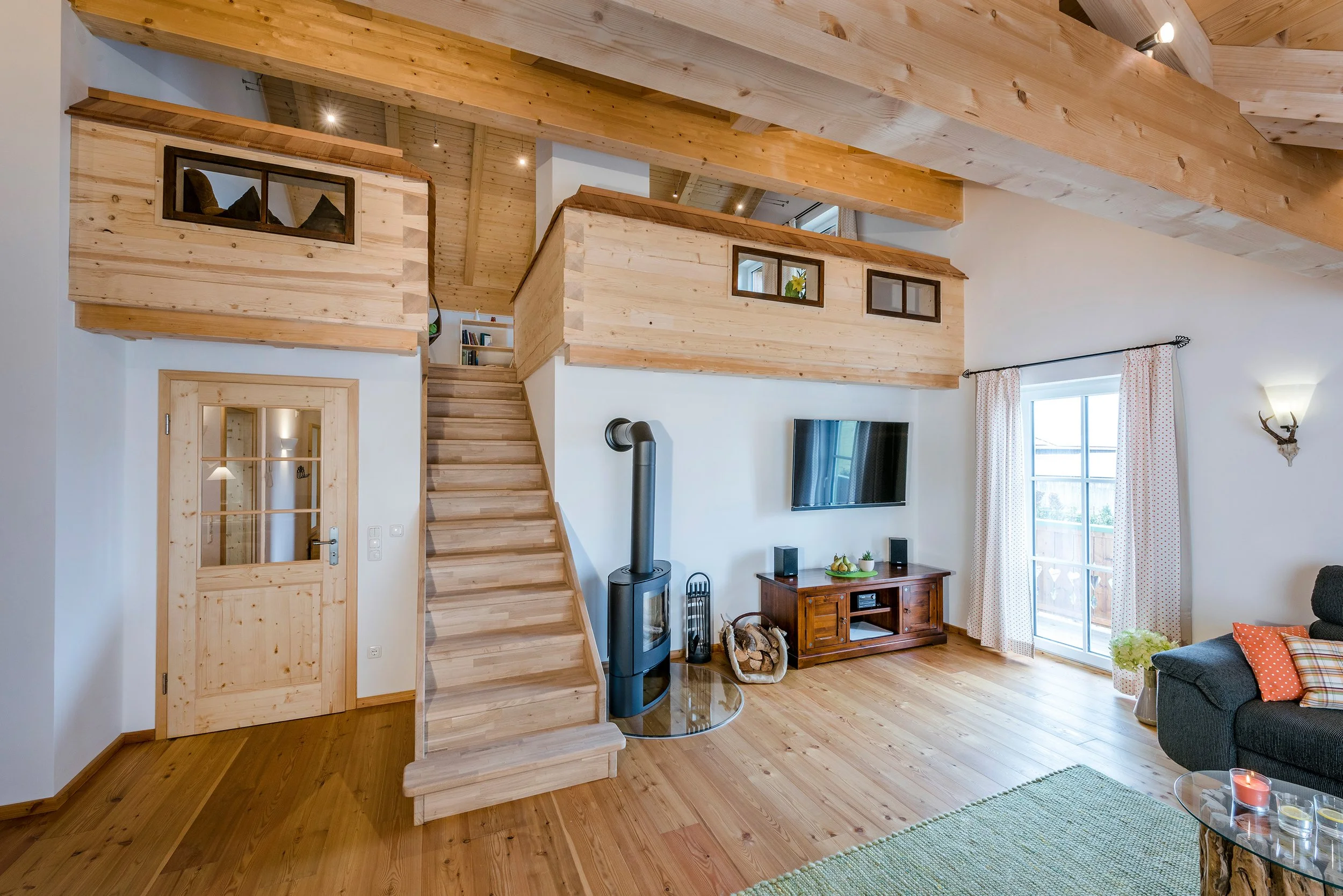 Wohnzimmer mit viel Holzdekor, offener Treppe, Kamin, Fernseher und großen Fenstern mit Gardinen.