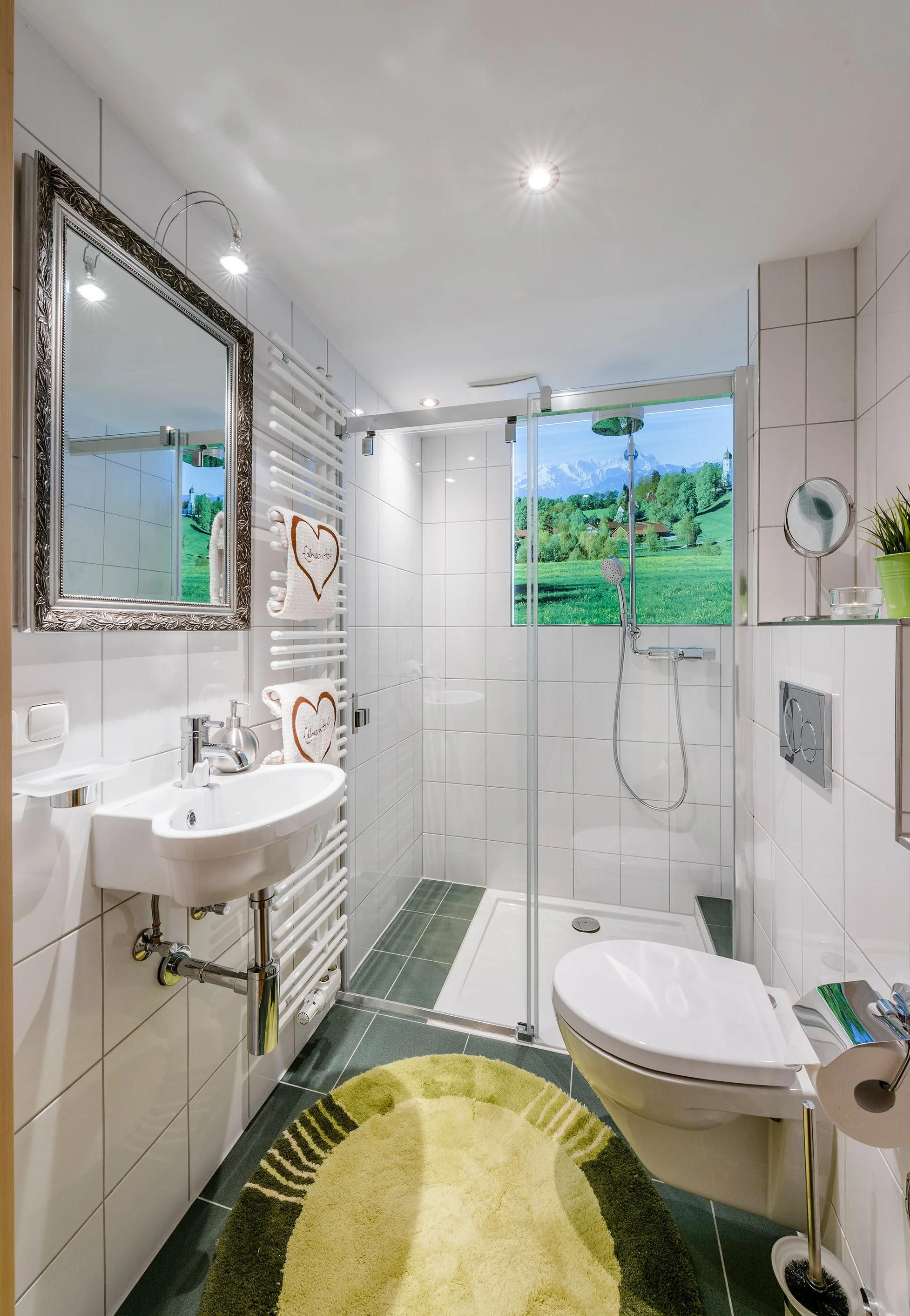 Ein modernes Badezimmer mit weiß gefliesten Wänden, einer Dusche mit Glaswand, einem weißen Becken, einem großen Spiegel mit dekorativem Rahmen und einem Fenster mit Blick auf grüne Wiesen und Berge. Es gibt eine grüne Badmatte auf dem Boden.