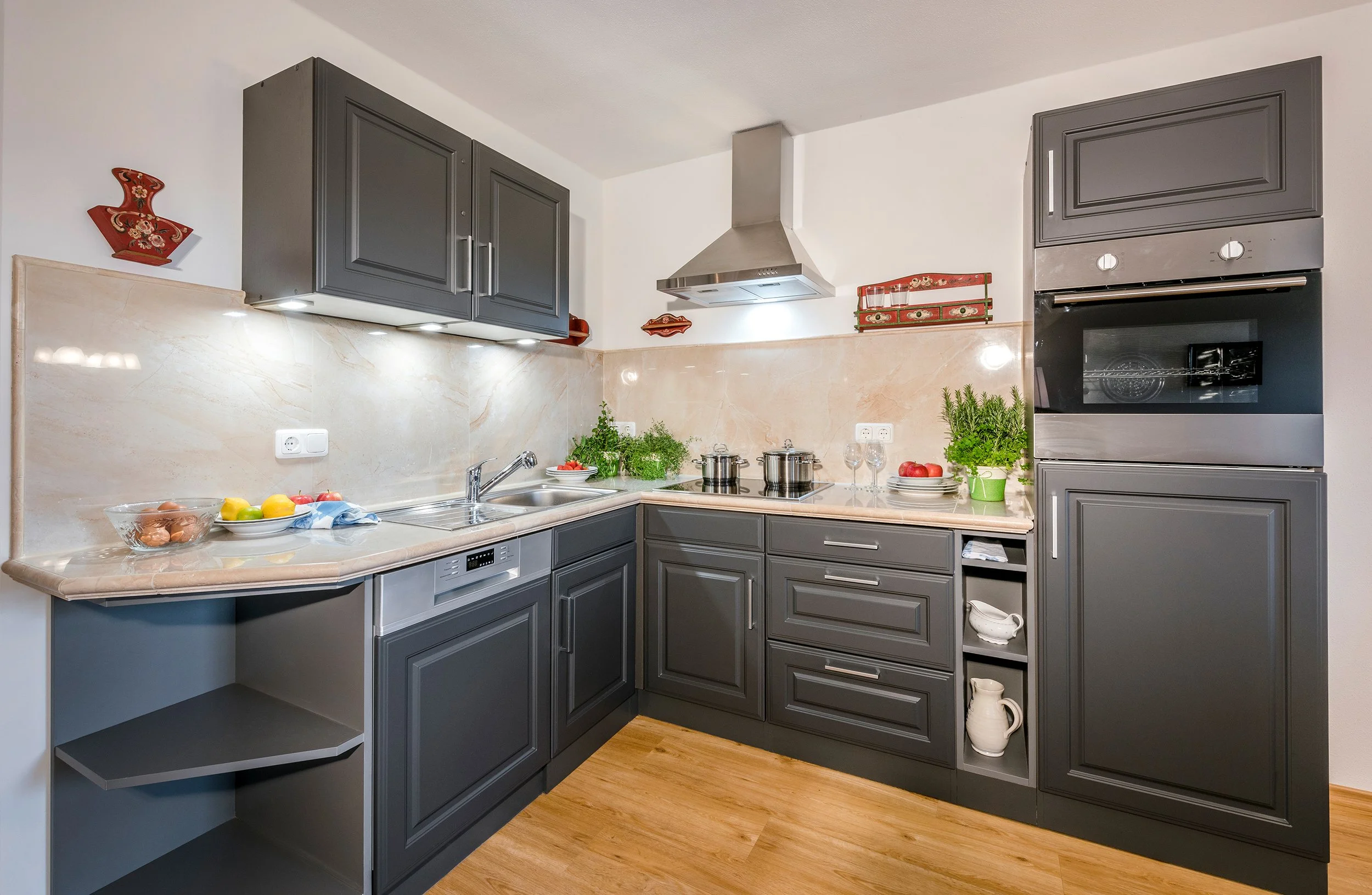 Moderne Küche mit grauen Schränken, Herd, Backofen, Spüle, Kochutensilien, frischen Kräutern, Obst und Dekorationen.