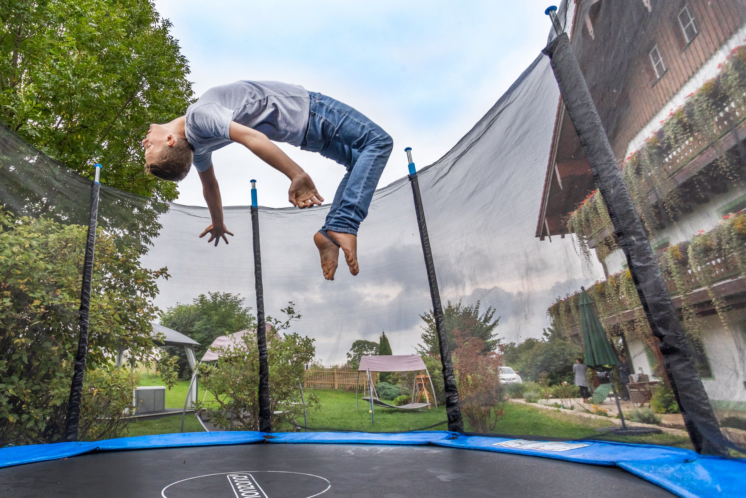 Ein Junge springt auf einem Trampolin im Garten, umgeben von Bäumen und einem Haus im Hintergrund.