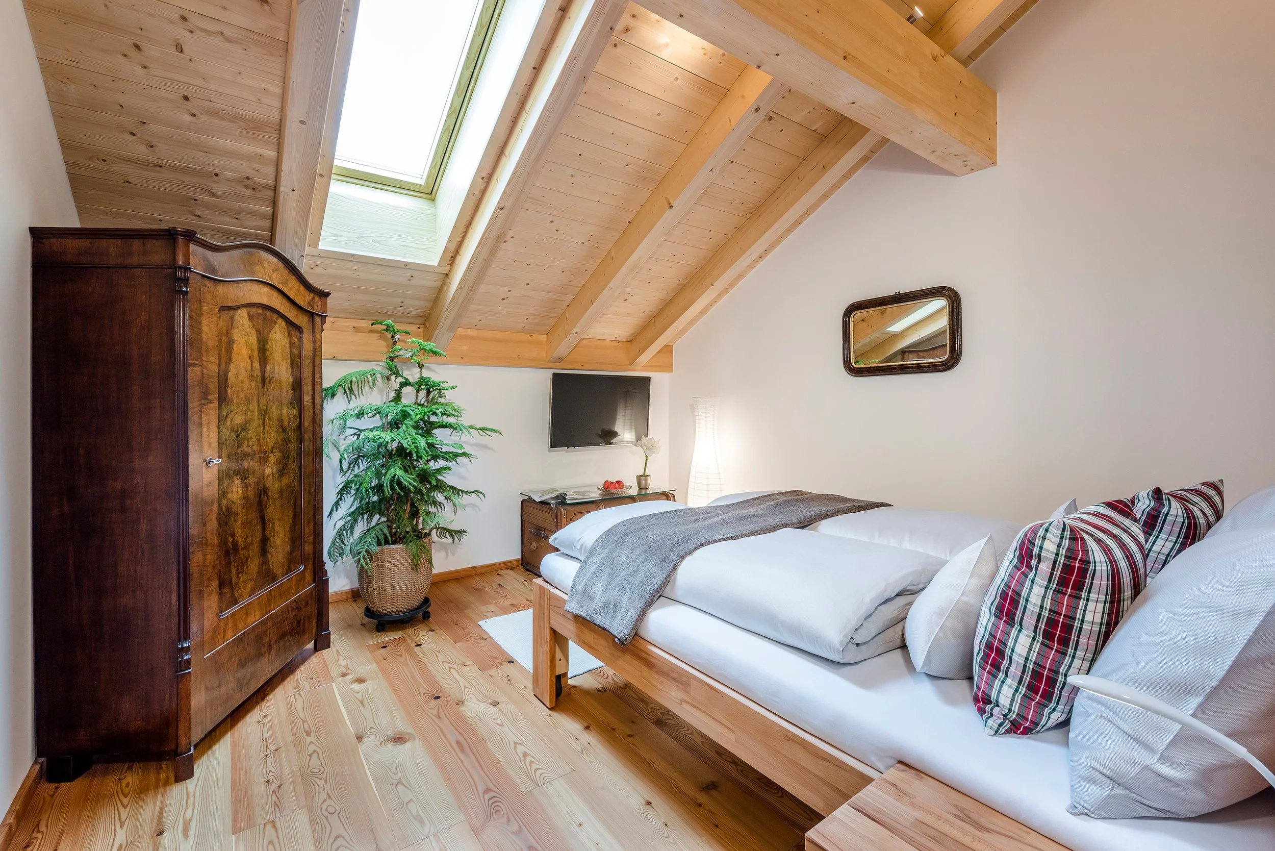 Helles Schlafzimmer mit Holzbalkendecke, Dachfenster, Doppelbett, antikem Kleiderschrank und Zimmerpflanze
