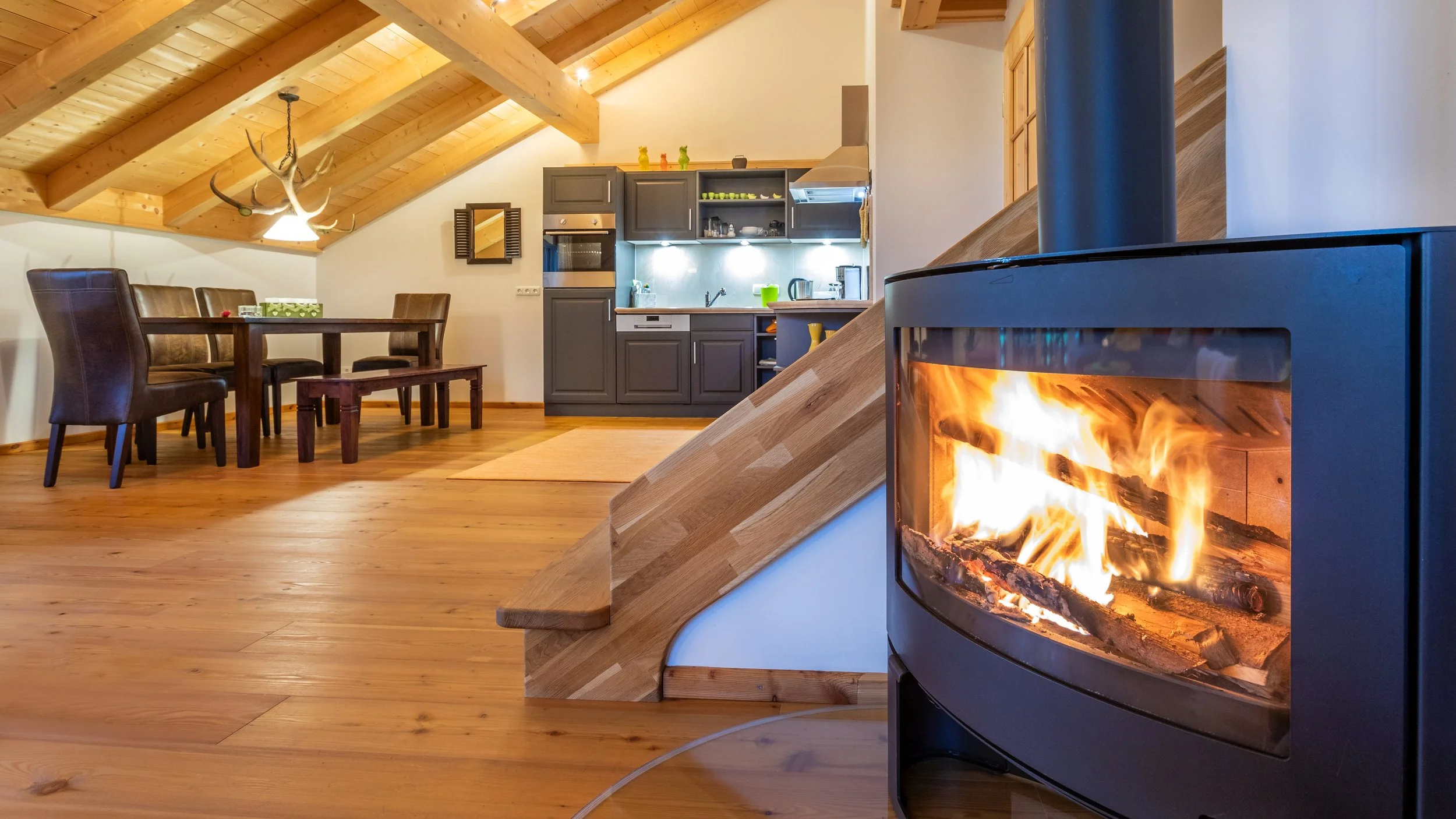 Gemütliches Wohnzimmer mit Holzdach, Kamin mit Feuer, Esstisch mit Stühlen und moderne Küchenzeile.