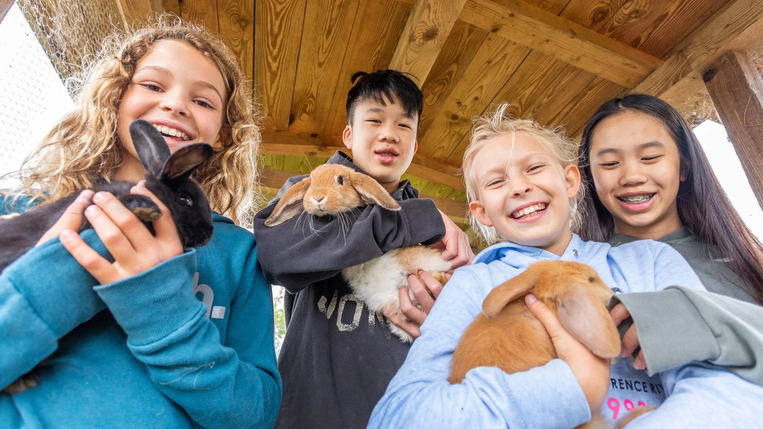 Fünf Kinder halten Hasen in den Armen, lachen und schauen in die Kamera, unter einem Holzüberdach.
