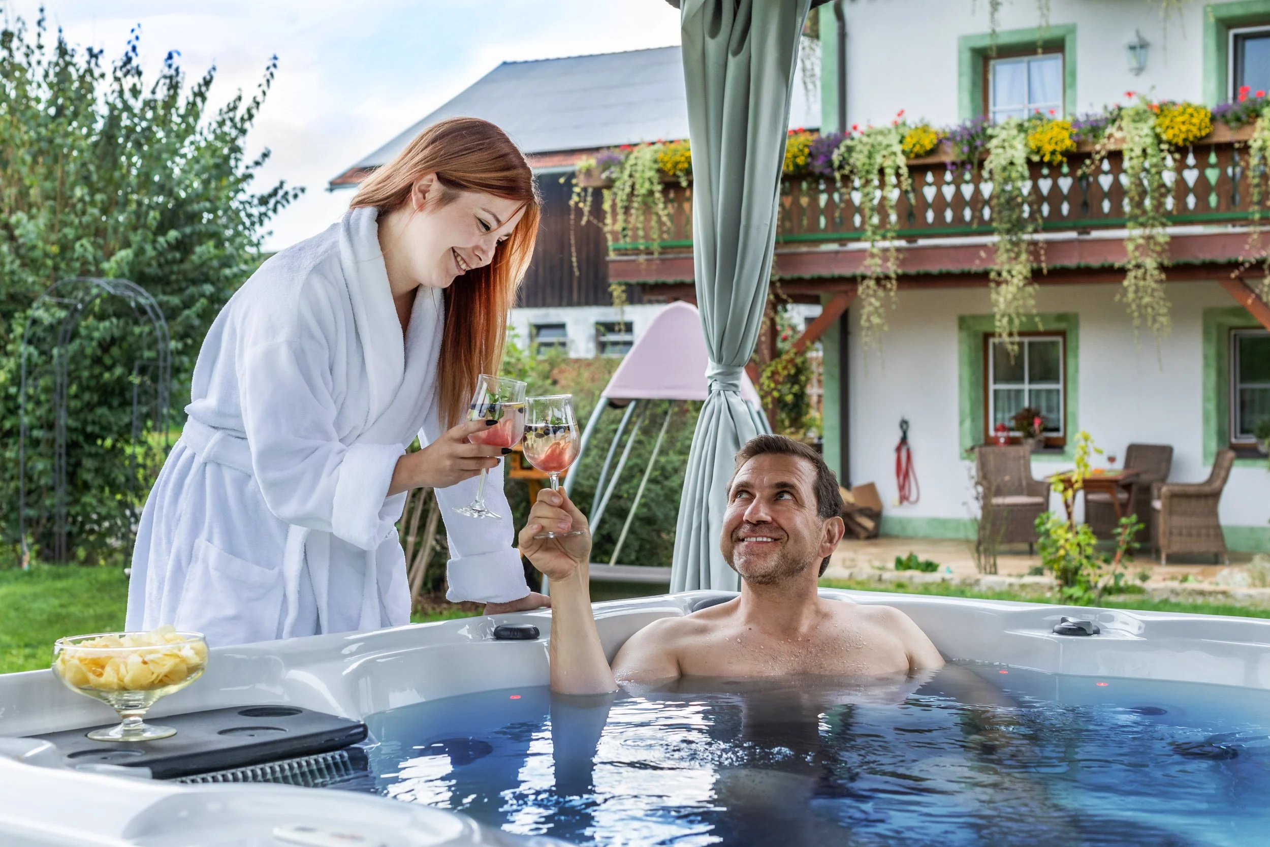 Frau hilft Mann im Whirlpool mit einem Glas Wein beim Anstoßen, im Hintergrund ein Haus mit Balkon und Blumen, es entsteht eine entspannte Atmosphäre an einem sonnigen Tag im Garten.