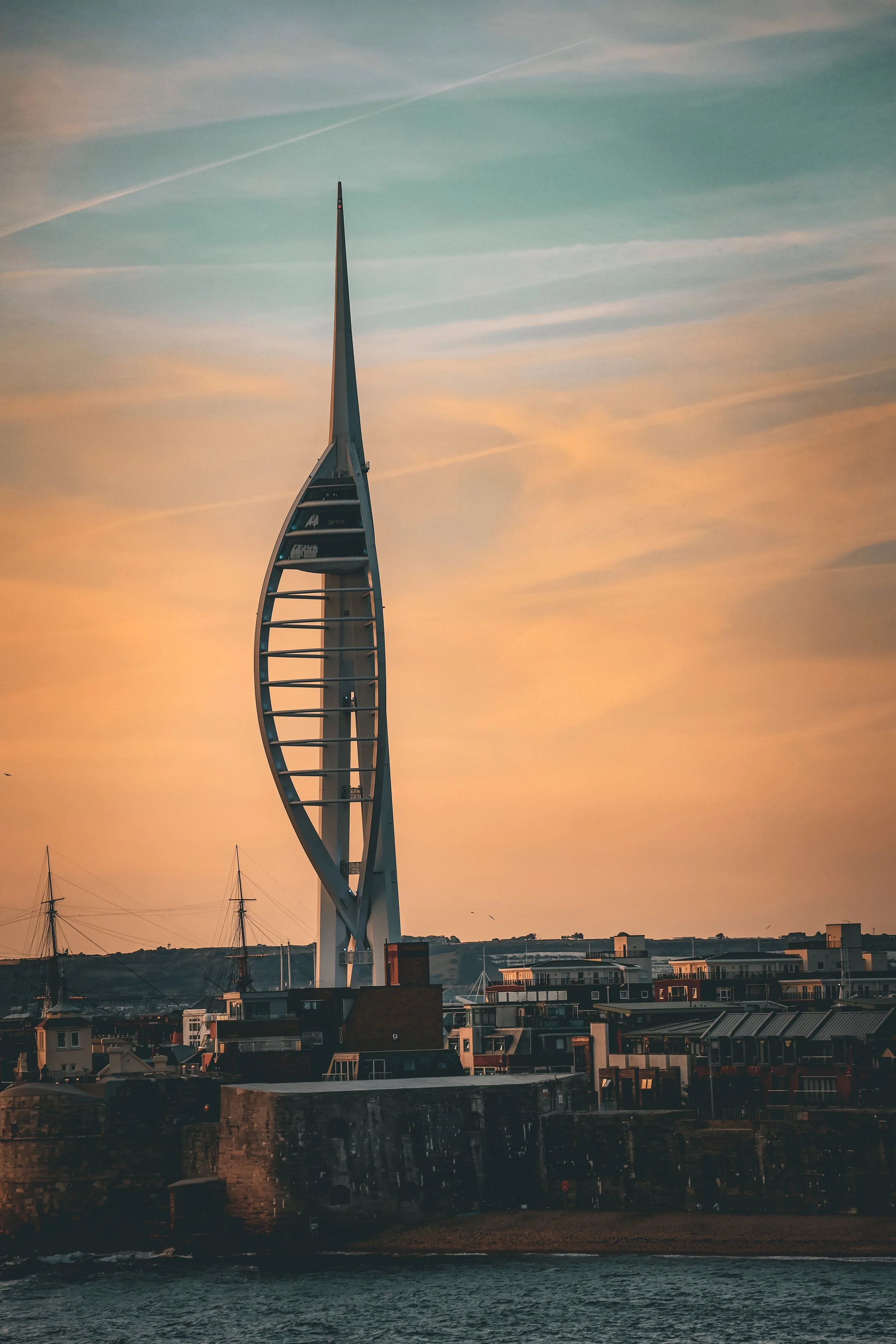 A modern architectural tower with a sail-like shape against a sunset sky, with sailing ships and city buildings in the foreground.