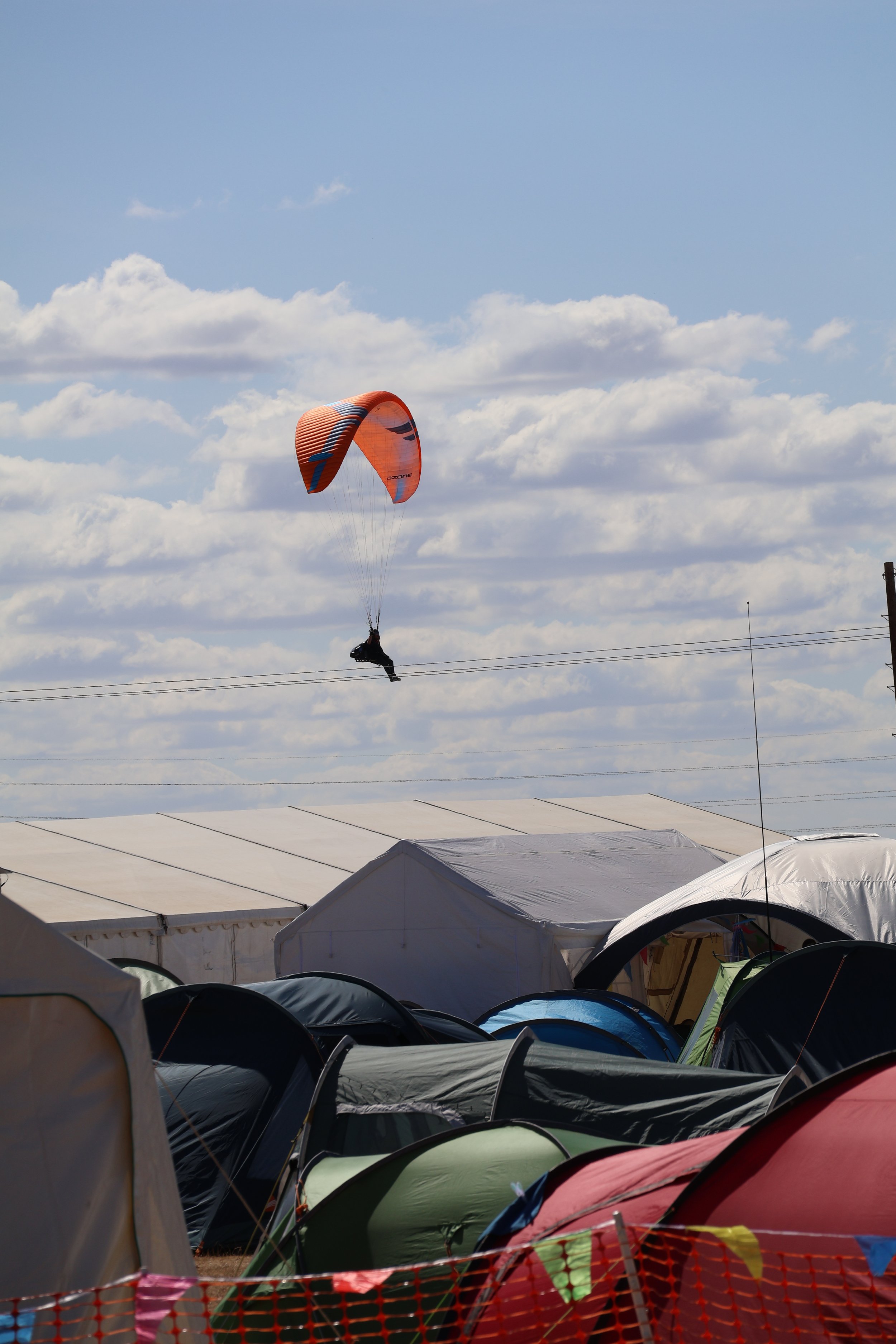 A man hang gliding under an orange and black parachute in a partly cloudy sky over a crowded outdoor camping area with various tents.
