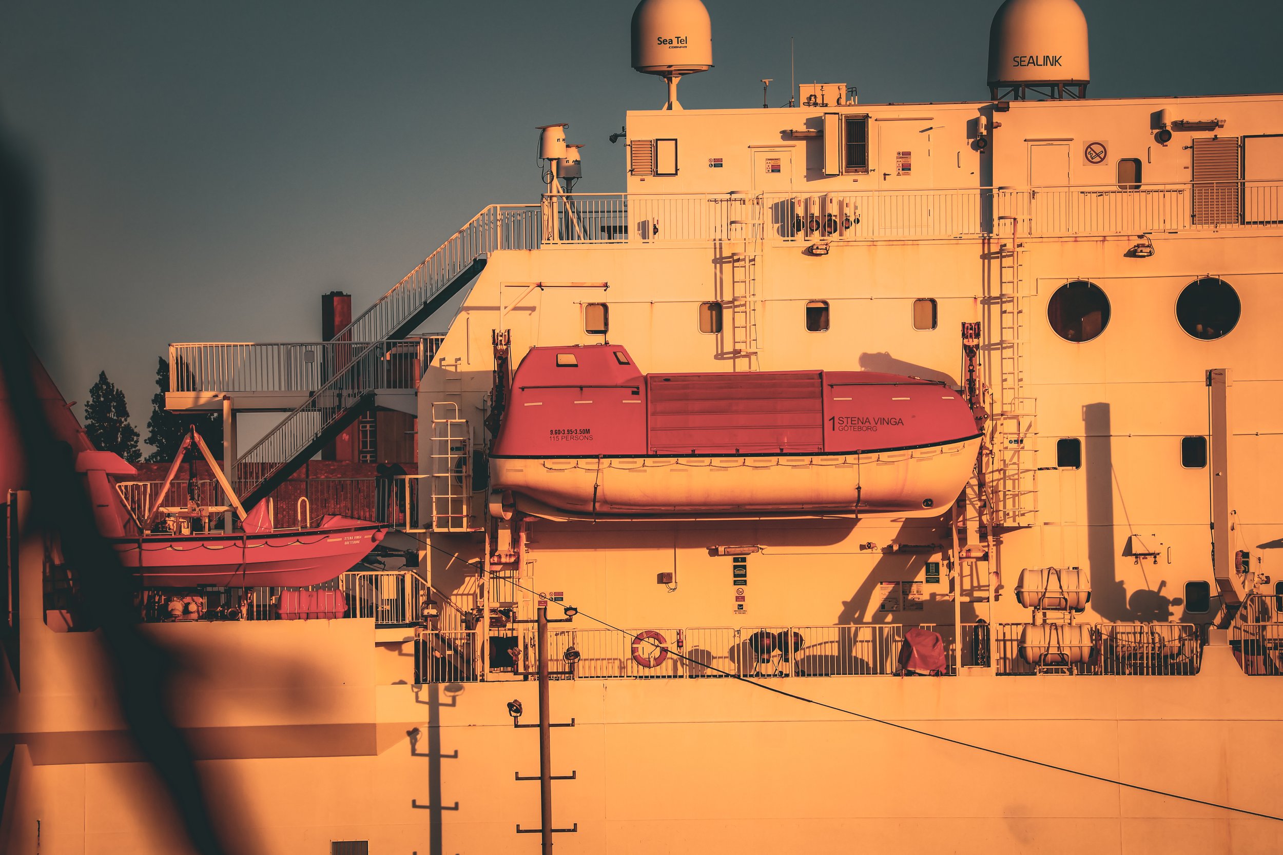 Close-up of the side of a large ship, illuminated by warm sunlight, featuring lifeboats, safety equipment, and communication antennas.
