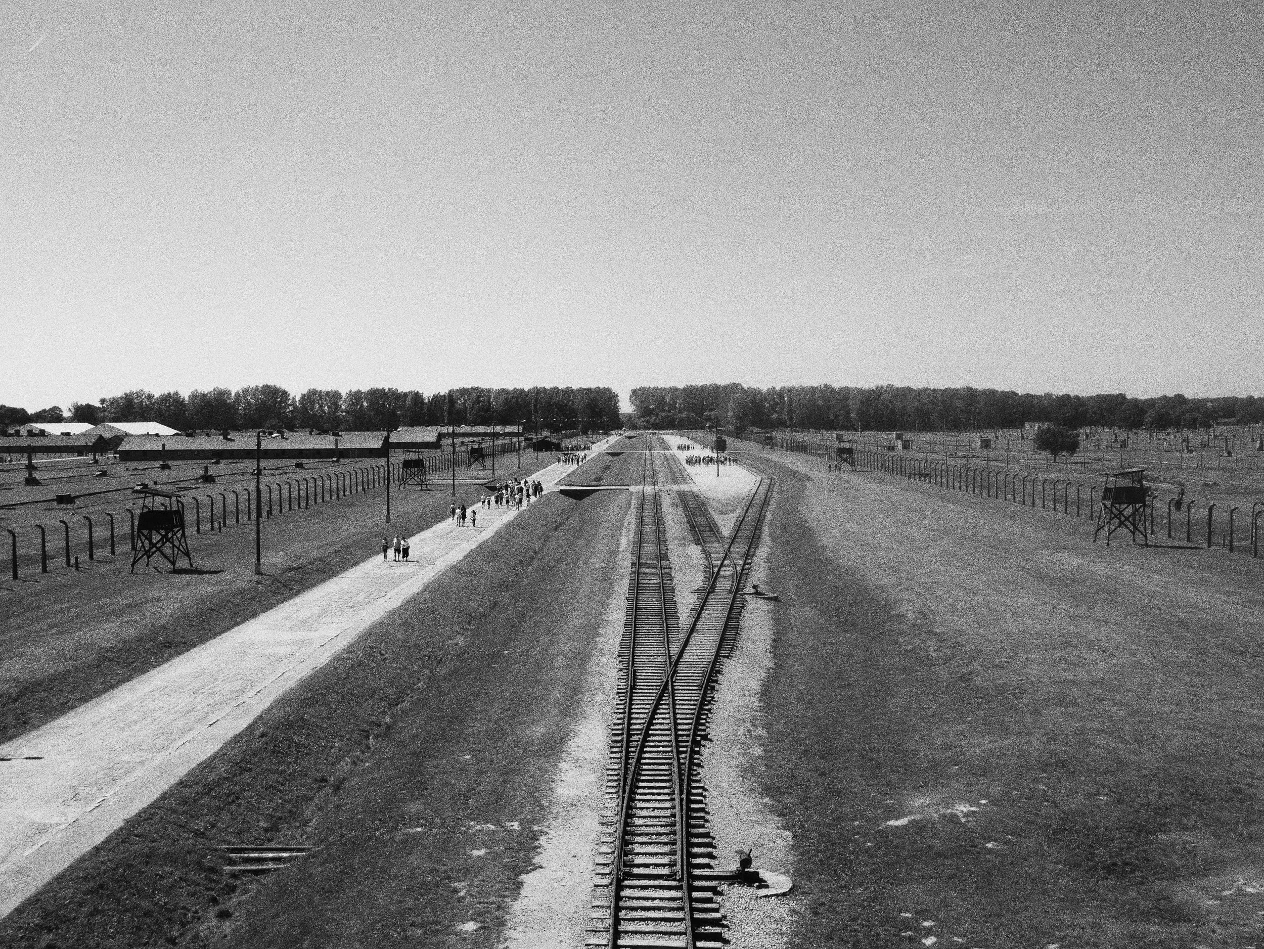 A black and white photo of a historical concentration camp with railway tracks, guard posts, and barbed wire fences, with a clear sky above and trees in the background.