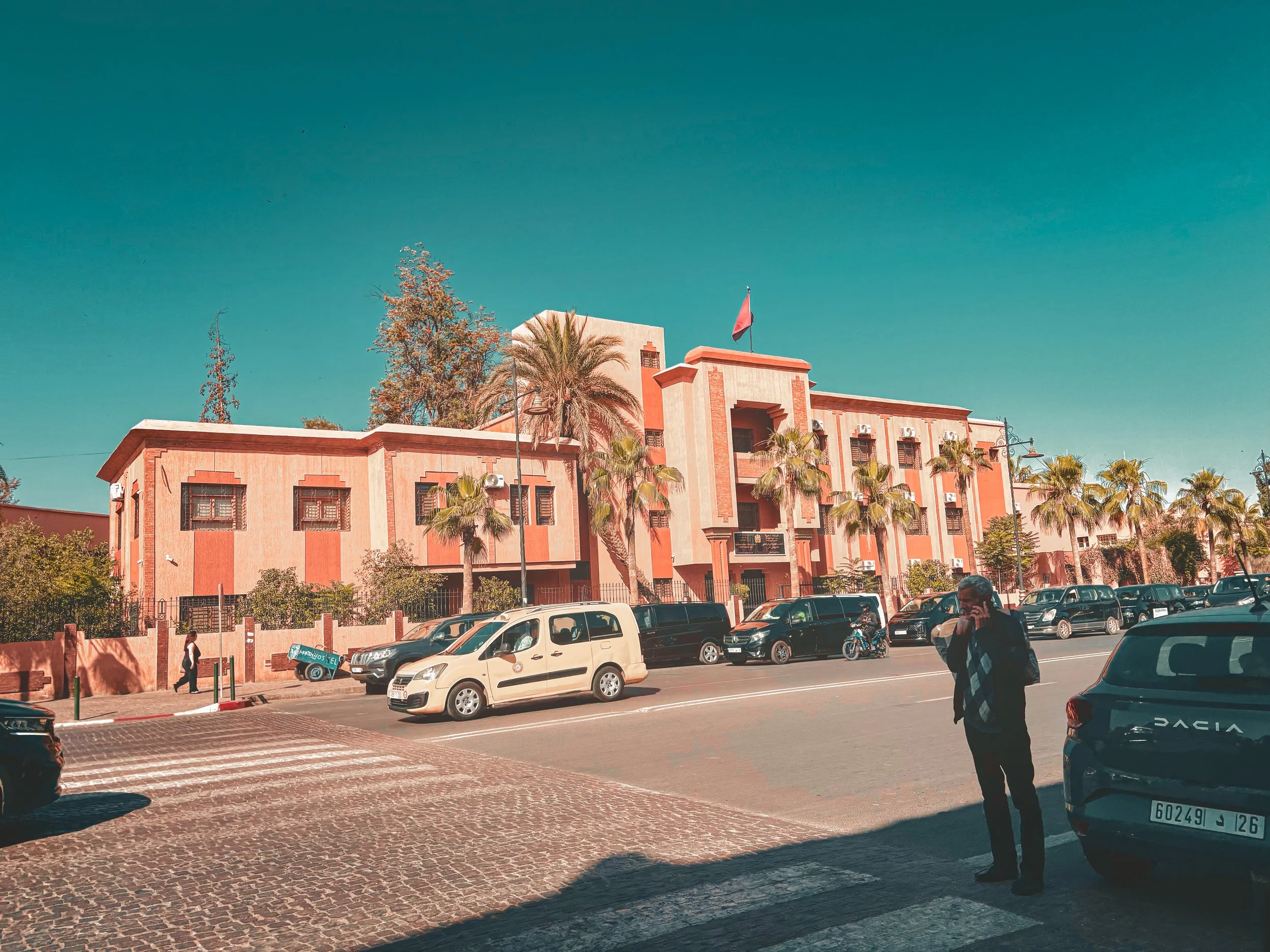 Street view with parked cars, a pink building with a flag on top, trees, and a person walking on the sidewalk.