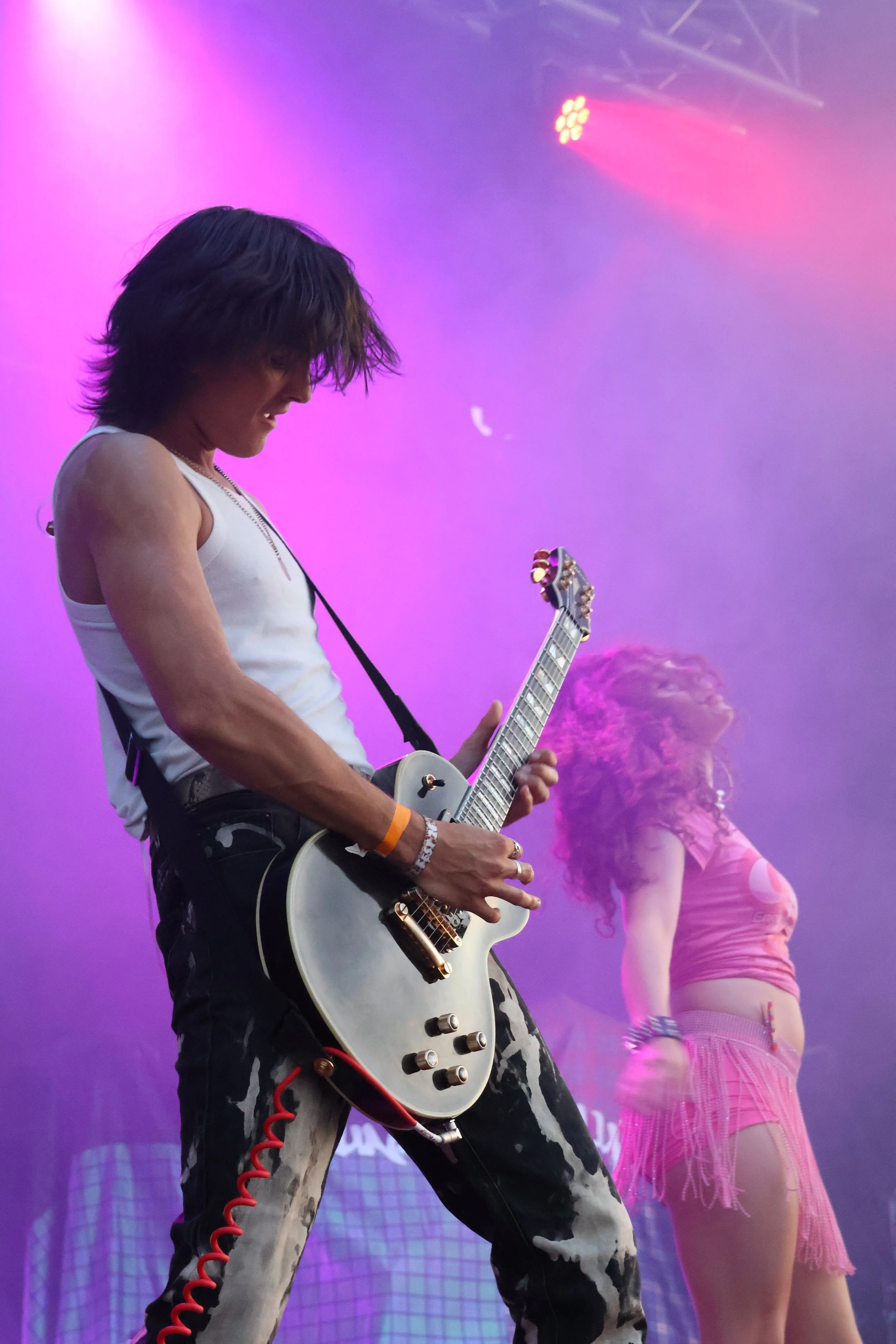 A male musician with long hair playing an electric guitar on a stage with purple and pink lighting. Behind him, a female dancer with curly hair and a pink outfit is visible.