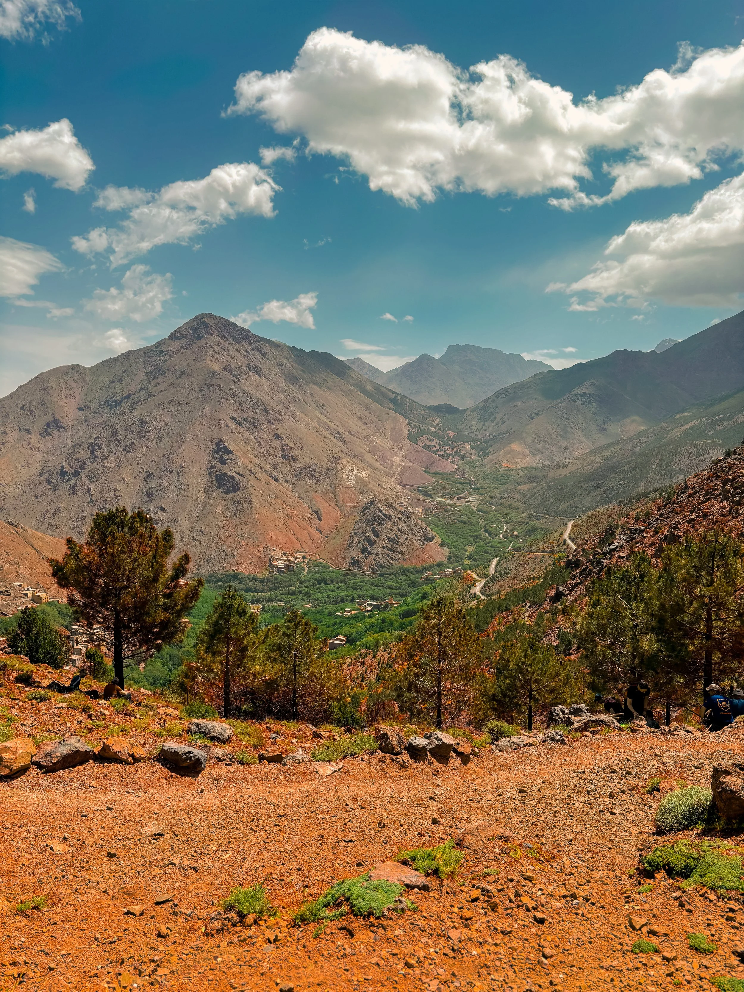 A mountainous landscape with rugged slopes, green valleys, scattered trees, and a partly cloudy sky.
