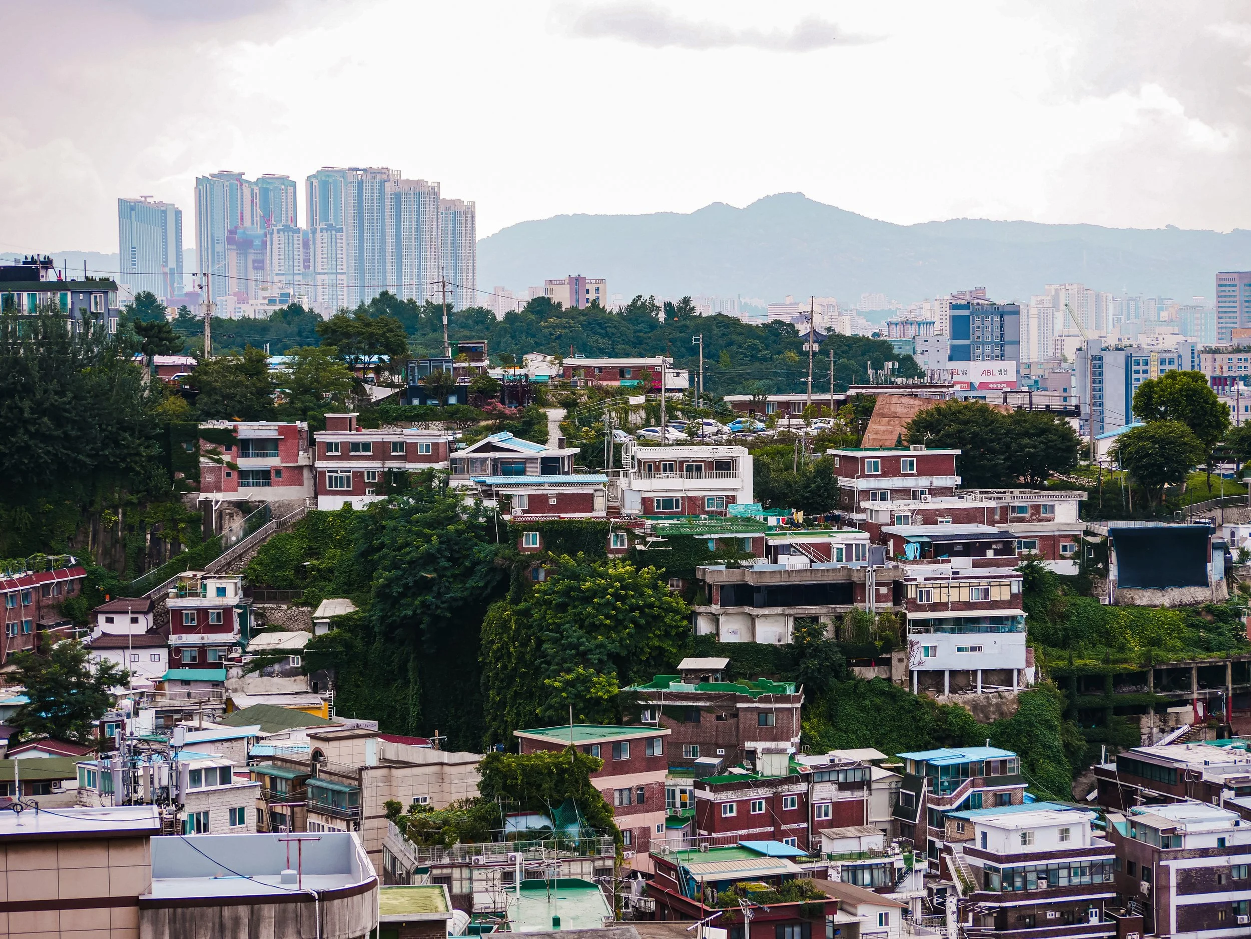 Cityscape with a mix of high-rise buildings and smaller residential houses, set against a mountain range in the background, under a cloudy sky.