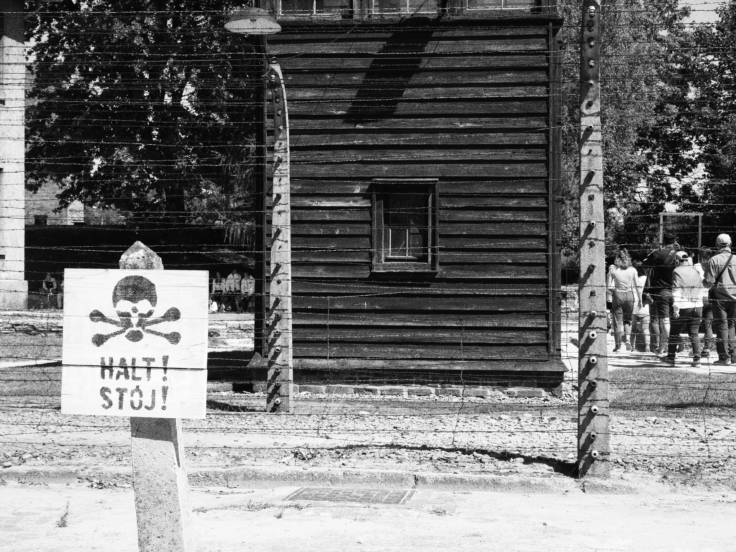 A locked wooden gate with barbed wire on top, surrounded by trees and people standing in a group in the background. There is a white sign with a skull and crossbones and Polish text that says 'Halt! Stój!' in front of the gate.