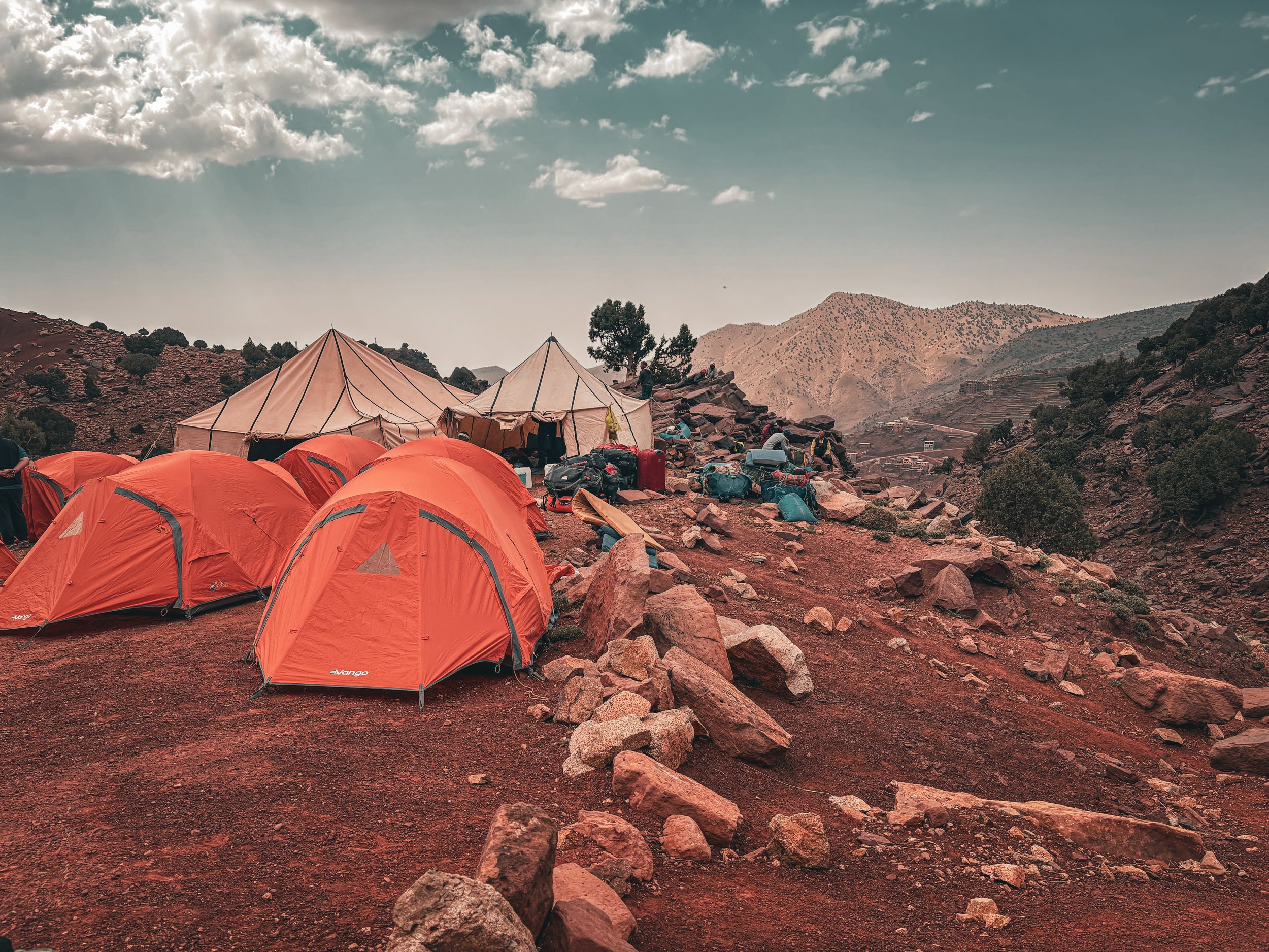 A landscape scene with orange tents set up on rocky terrain in a mountainous area with hills and sparse vegetation, under a partly cloudy sky.