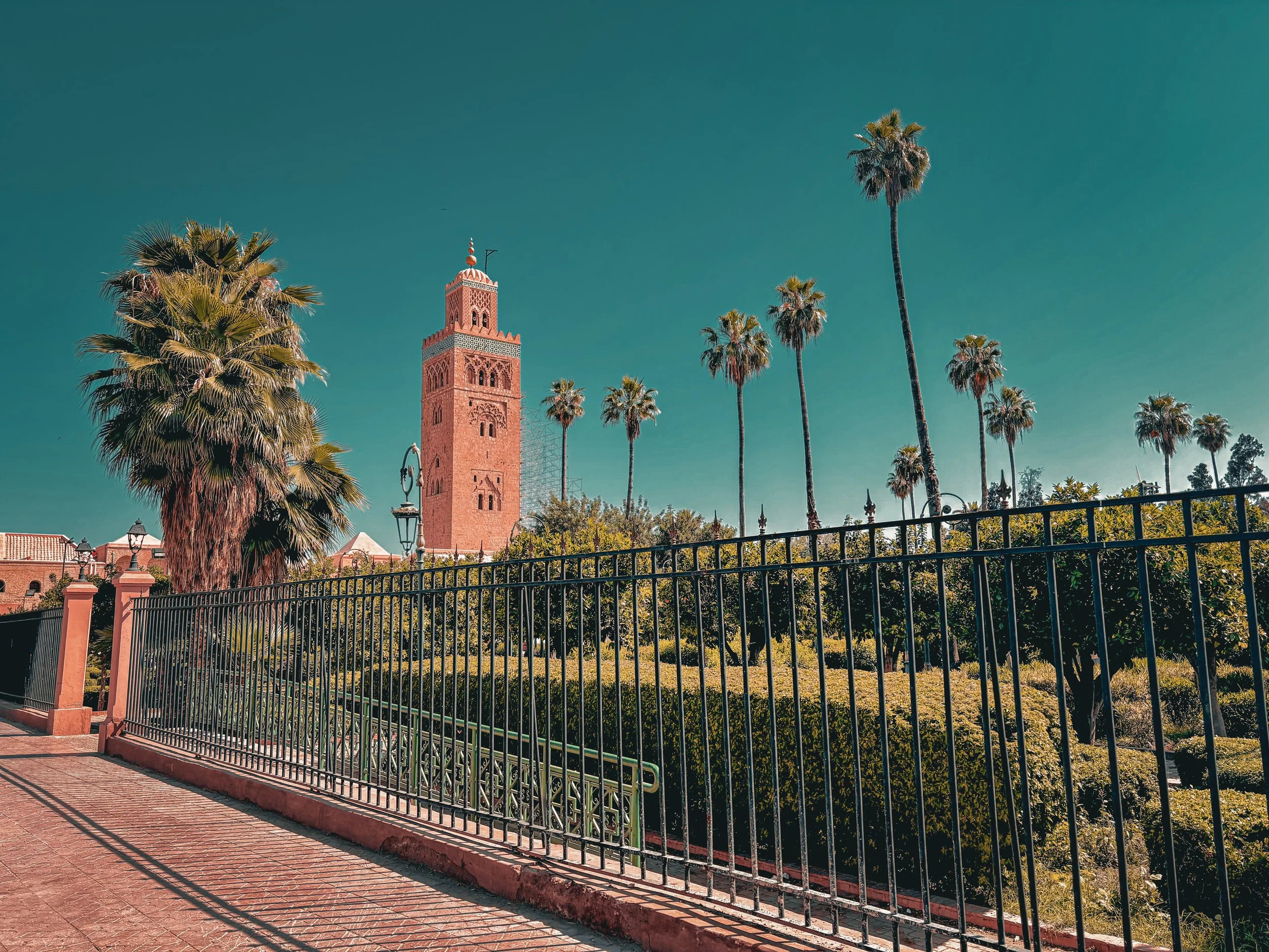 A scene of a city park with a black wrought iron fence, lush green bushes, and tall palm trees. Prominent in the background is a reddish tower with intricate architectural details, set against a clear blue sky with no clouds.