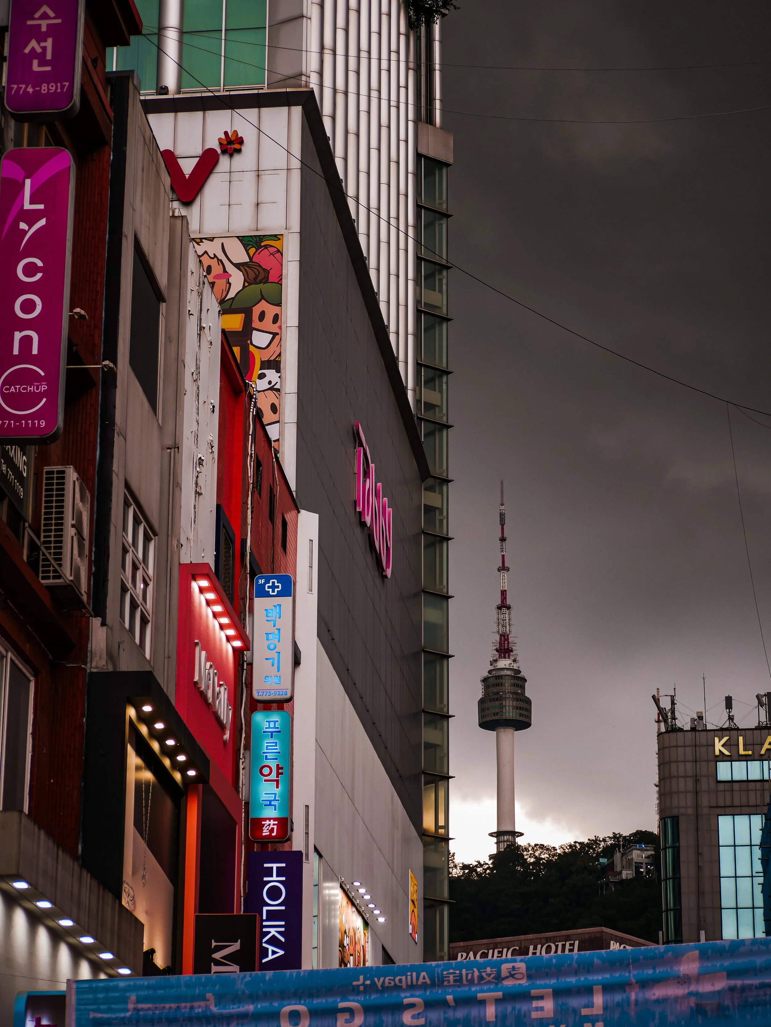 A city street scene in Seoul, South Korea, showing illuminated signage on buildings, including a pink sign for a cosmetics store, and a view of Namsan Tower and a television tower on a dark, cloudy sky in the distance.