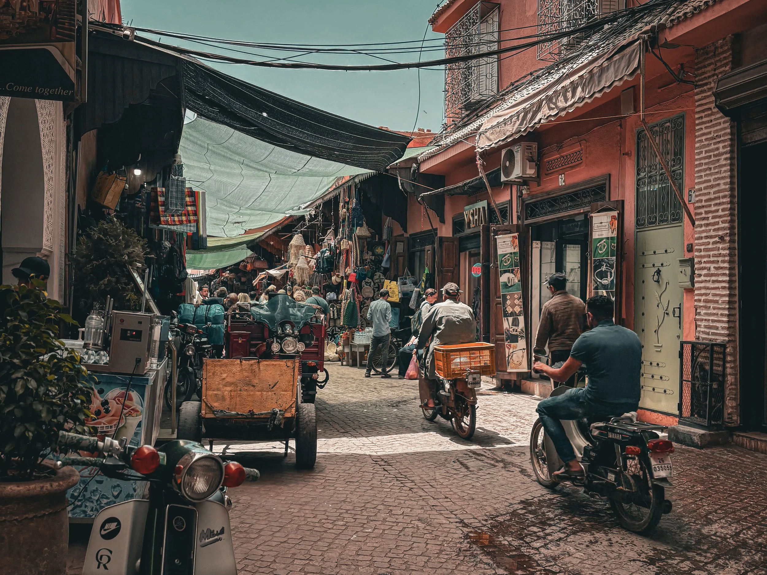 Street scene in a marketplace with shops, people walking, and motorbikes parked along a cobblestone street, with overhead canopies providing shade.