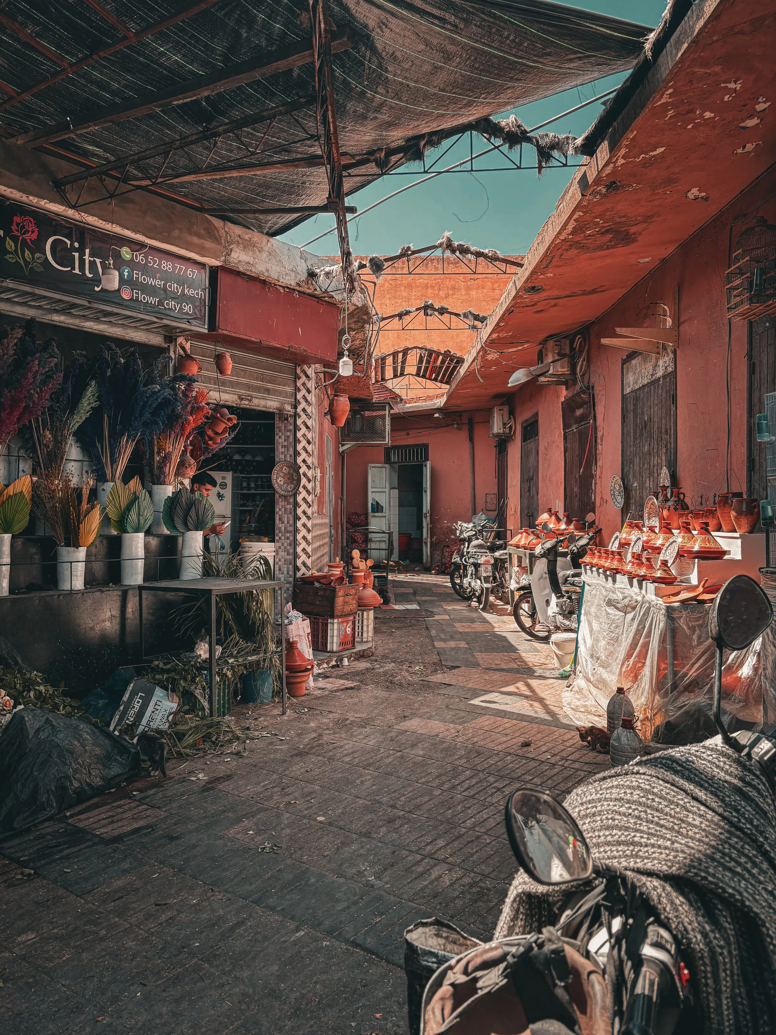 An outdoor marketplace or street scene with small shops and vendors. There are motorcycles and scooters parked along the narrow pathway. Items such as pottery and decorative objects are displayed outside the shops, and the area is covered by a makeshift roof made of fabric and poles. The buildings have a weathered appearance with reddish walls.